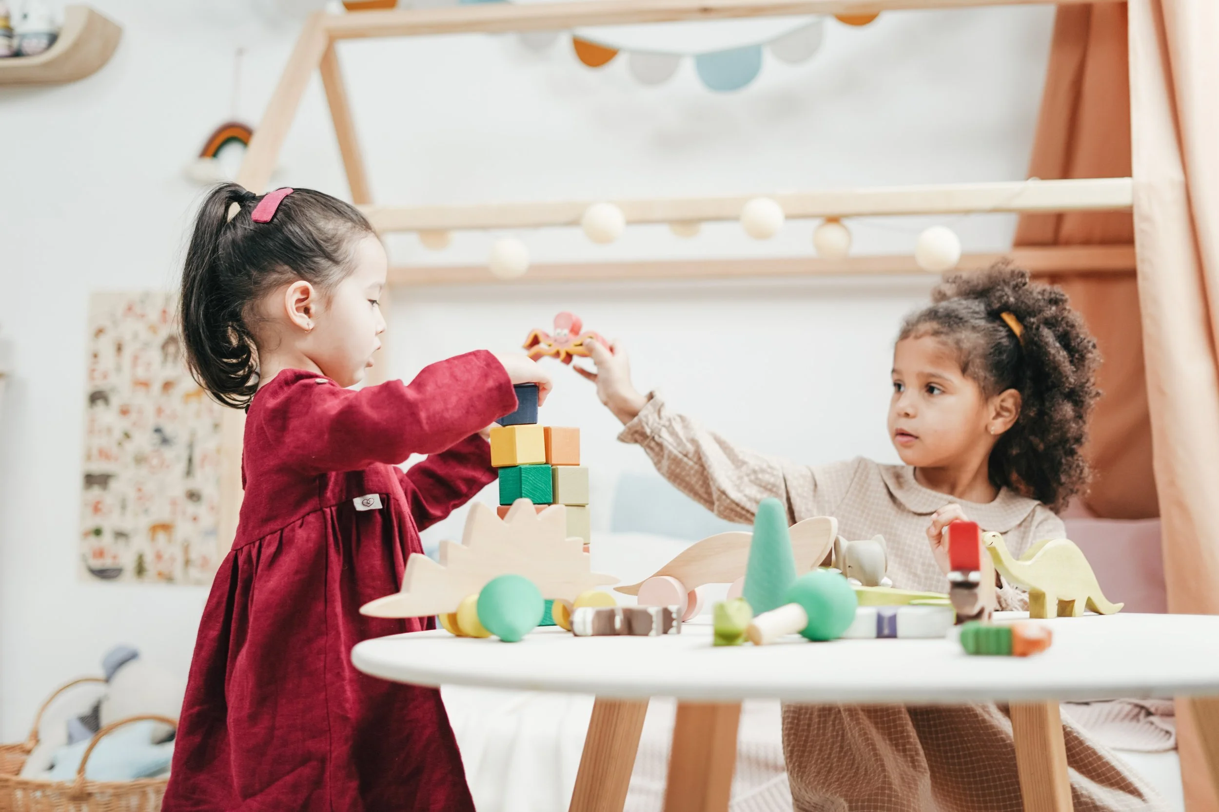 Two young girls playing with colorful wooden blocks and toys at a white table in a cozy, decorated room.