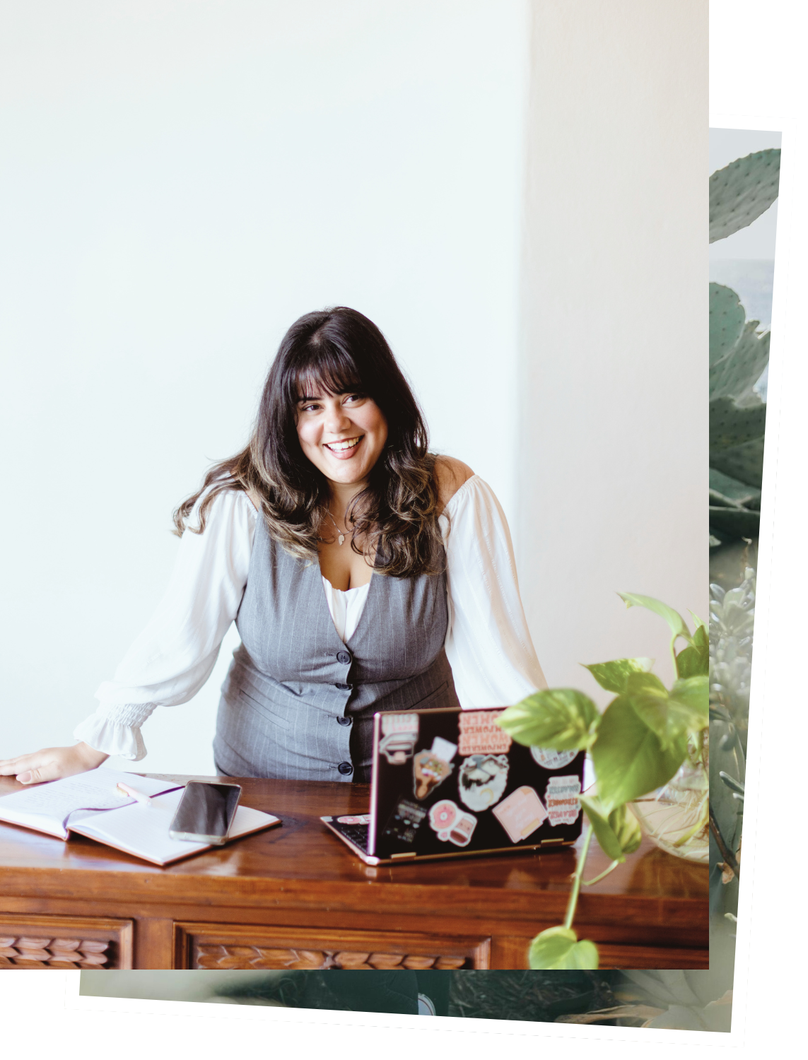 Smiling woman with dark hair in a gray vest and white blouse working at a wooden desk with a laptop, notebook, and smartphone, next to a green plant.