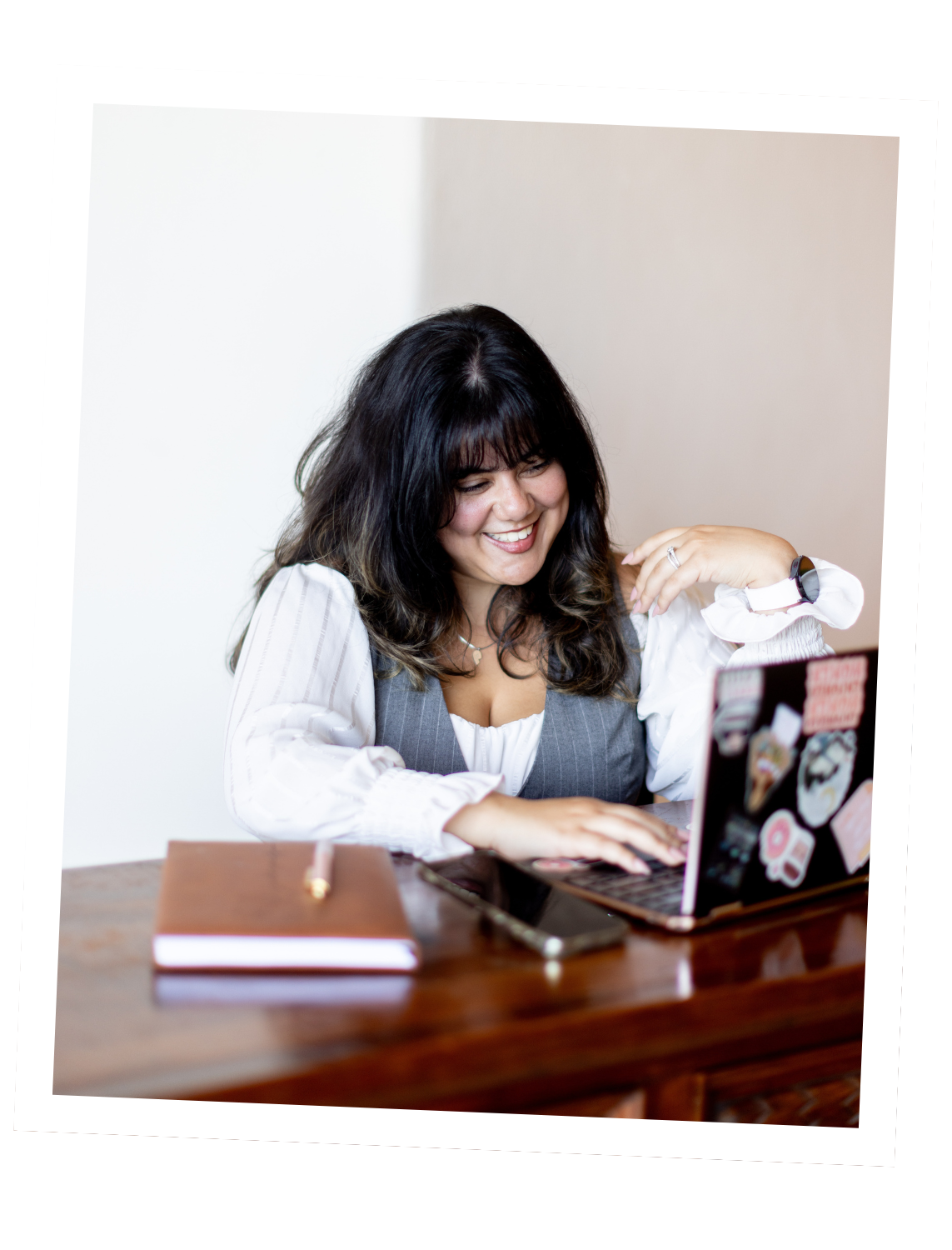 Woman with dark curly hair smiling while working on a laptop at a desk, with a notebook and pen nearby.