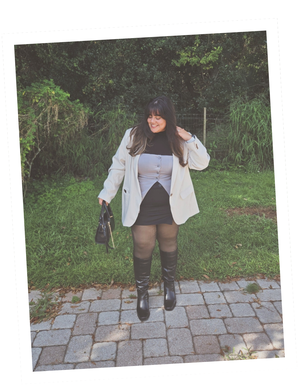 A woman standing outdoors on a paved path, smiling, with greenery and trees in the background, dressed in a beige blazer, gray top, black skirt, and black thigh-high boots, carrying a black handbag.