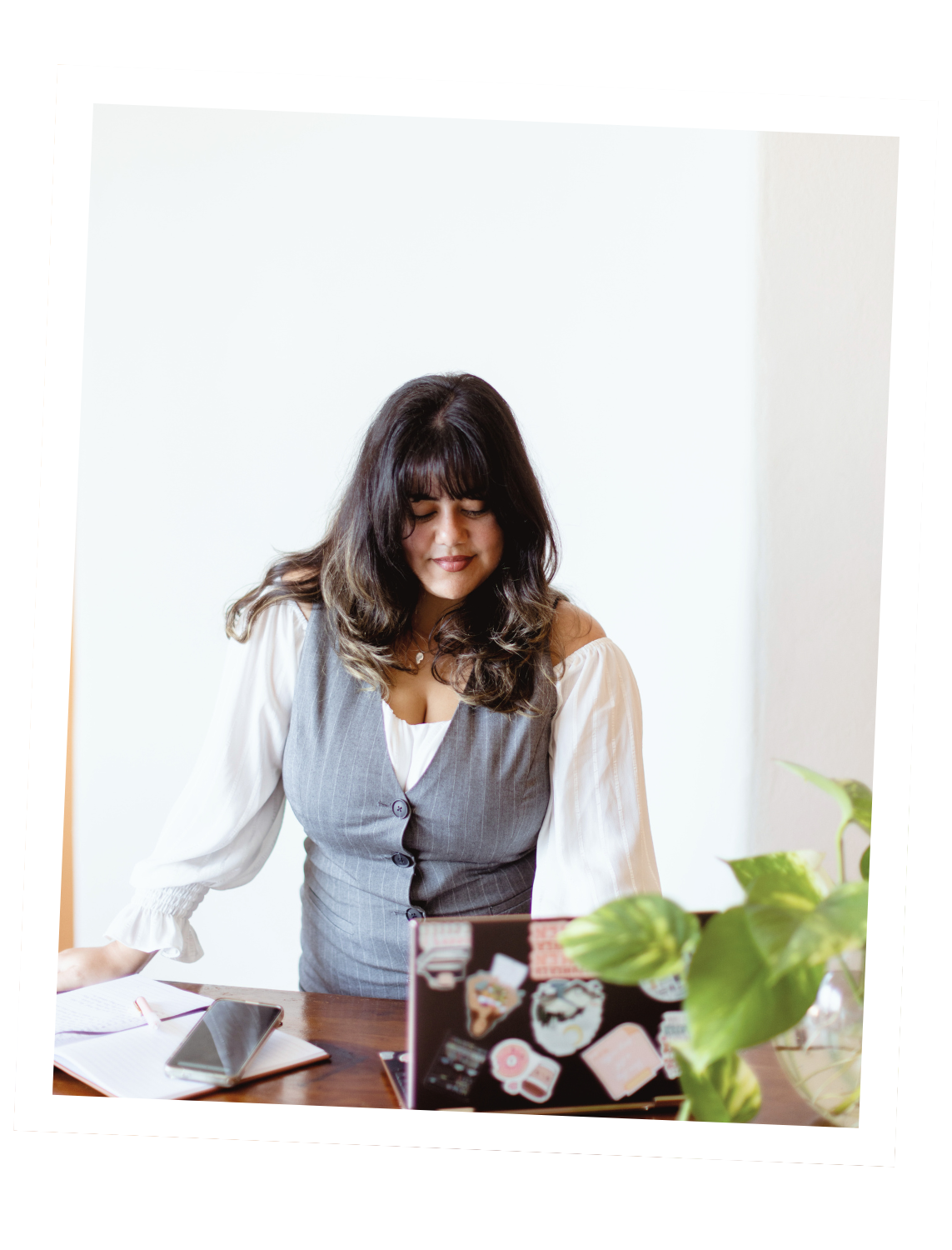 A woman with dark hair and bangs, wearing a white blouse and gray vest, standing at a table with a laptop covered in stickers, an open notebook, a smartphone, and a green potted plant nearby.