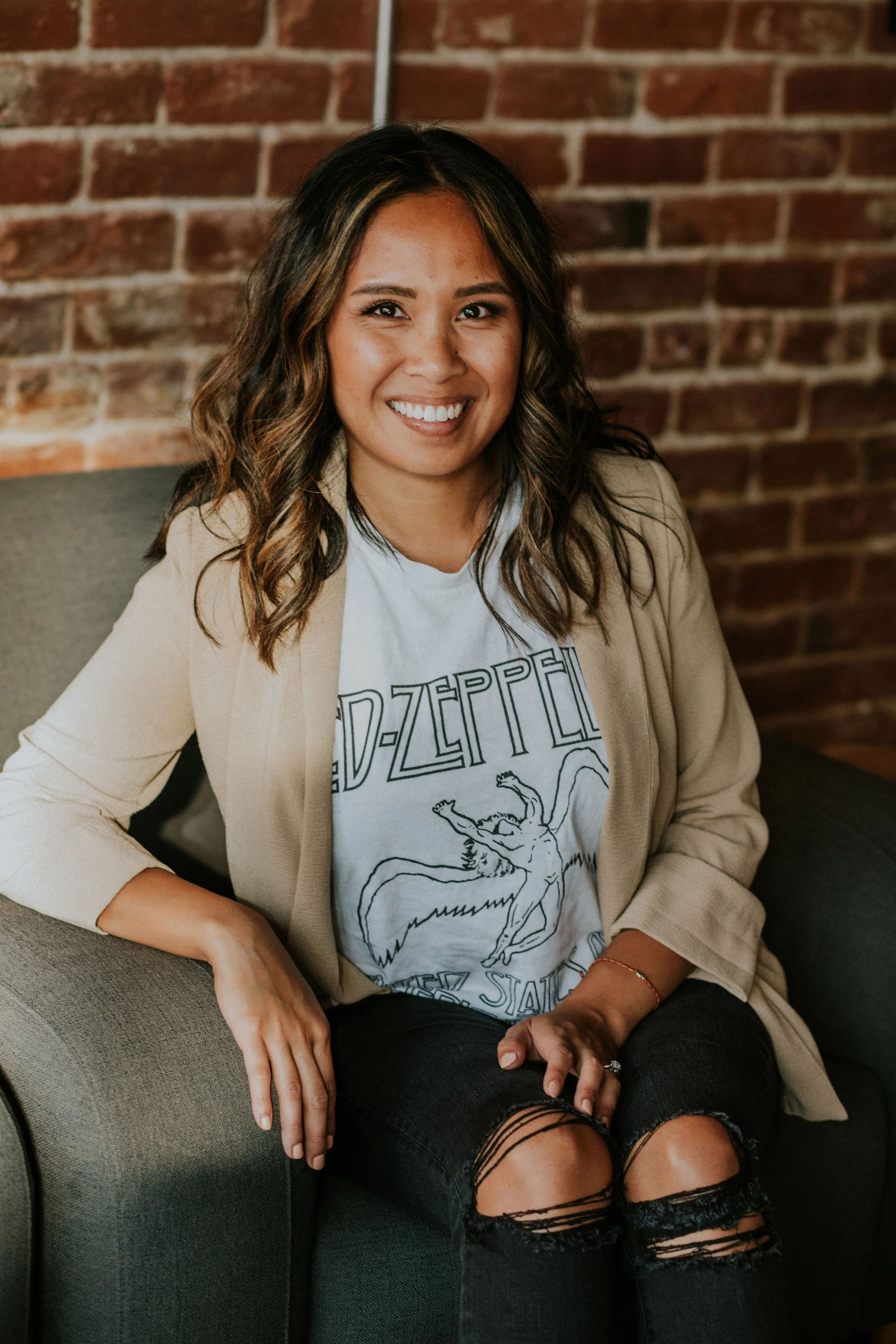 A woman sitting on a gray couch against a brick wall, smiling at the camera. She has wavy brown hair with highlights, is wearing a beige blazer, a white Led Zeppelin T-shirt, and ripped black jeans.