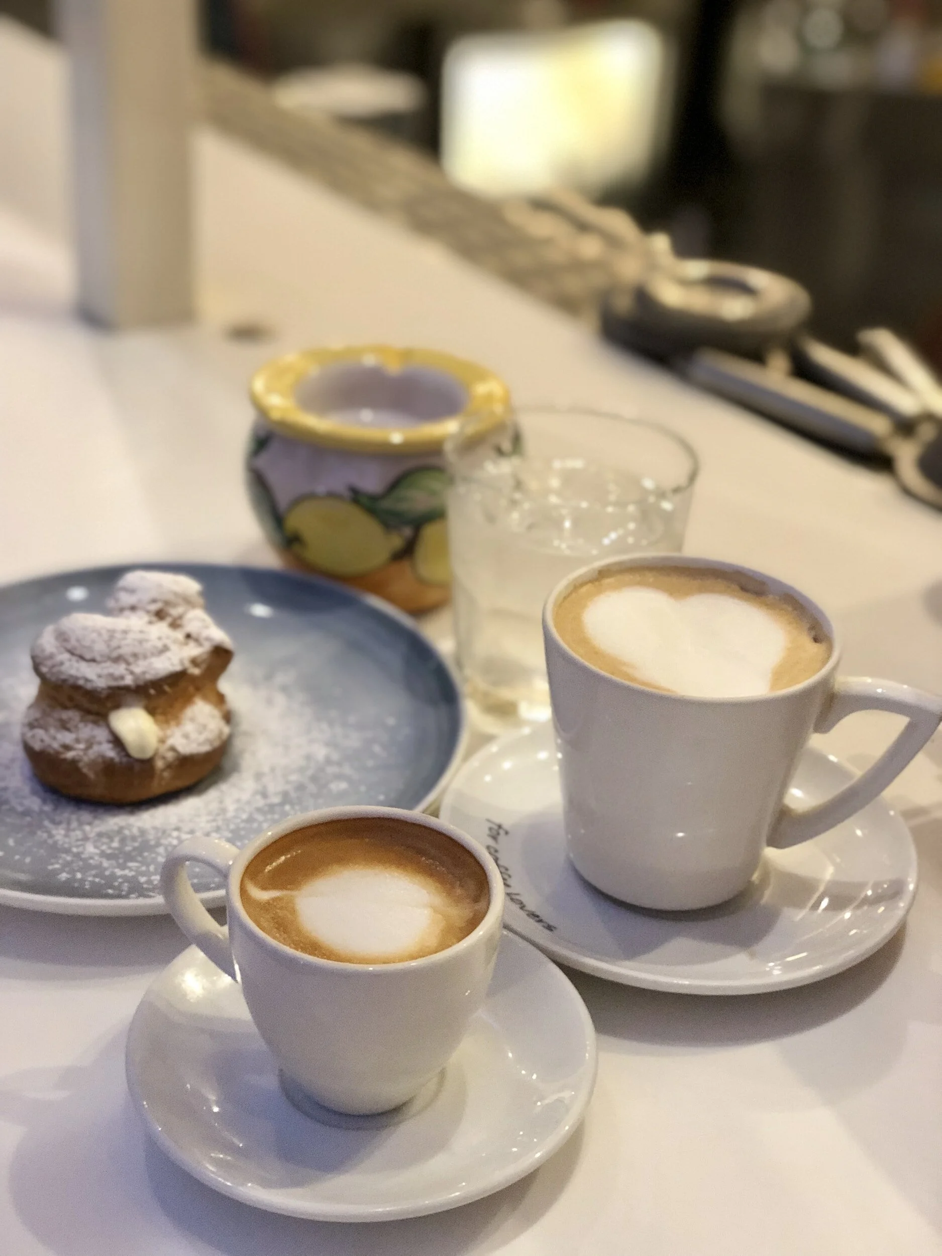 Two cups of cappuccino with foam art, a small plate with a cream-filled pastry dusted with powdered sugar, a glass of water, and a ceramic container with a lemon pattern on a white table.