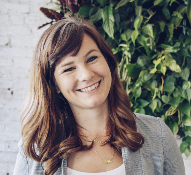 Smiling woman with brown hair and a gray blazer in front of a green leafy background.
