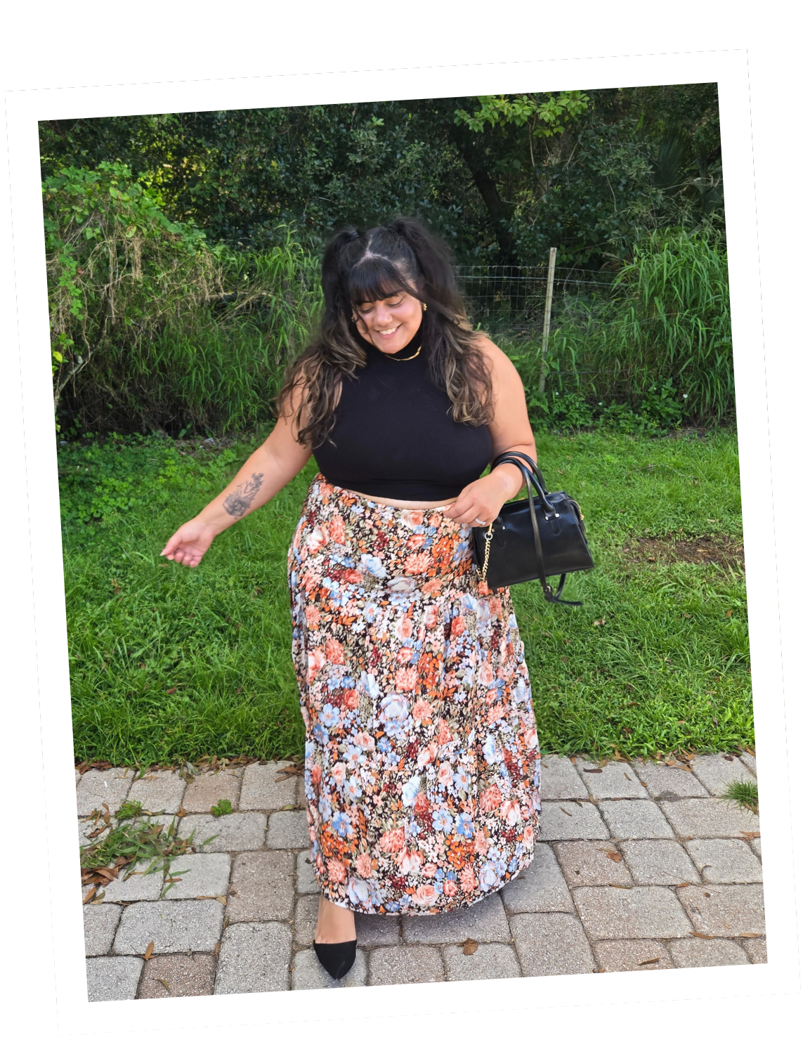 A woman with dark hair in pigtails, wearing a black sleeveless top, a long colorful floral skirt, black heels, and carrying a black handbag, standing on a brick pathway outdoors with green grass and bushes in the background, smiling.