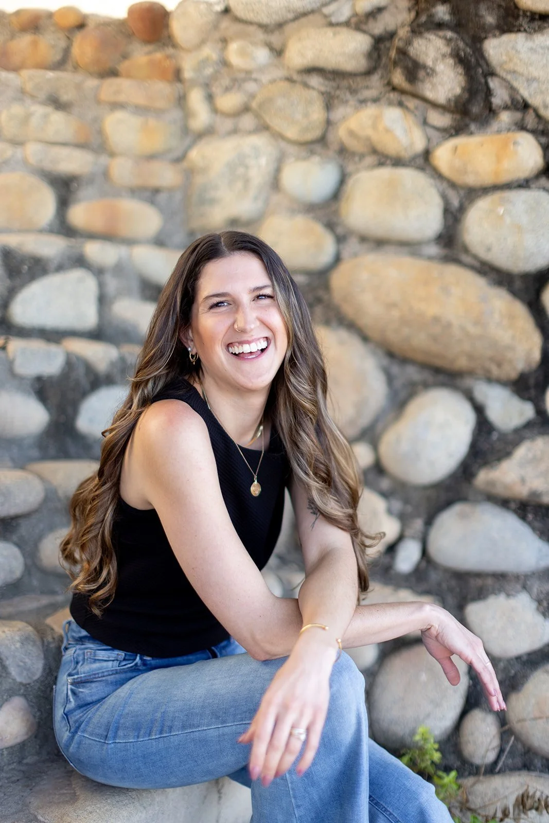 A woman with long wavy brown hair and light skin, smiling and laughing, sitting in front of a stone wall, wearing a black sleeveless top and blue jeans.