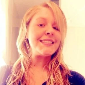 A young woman with long, wavy blonde hair smiling at the camera in a well-lit indoor setting.