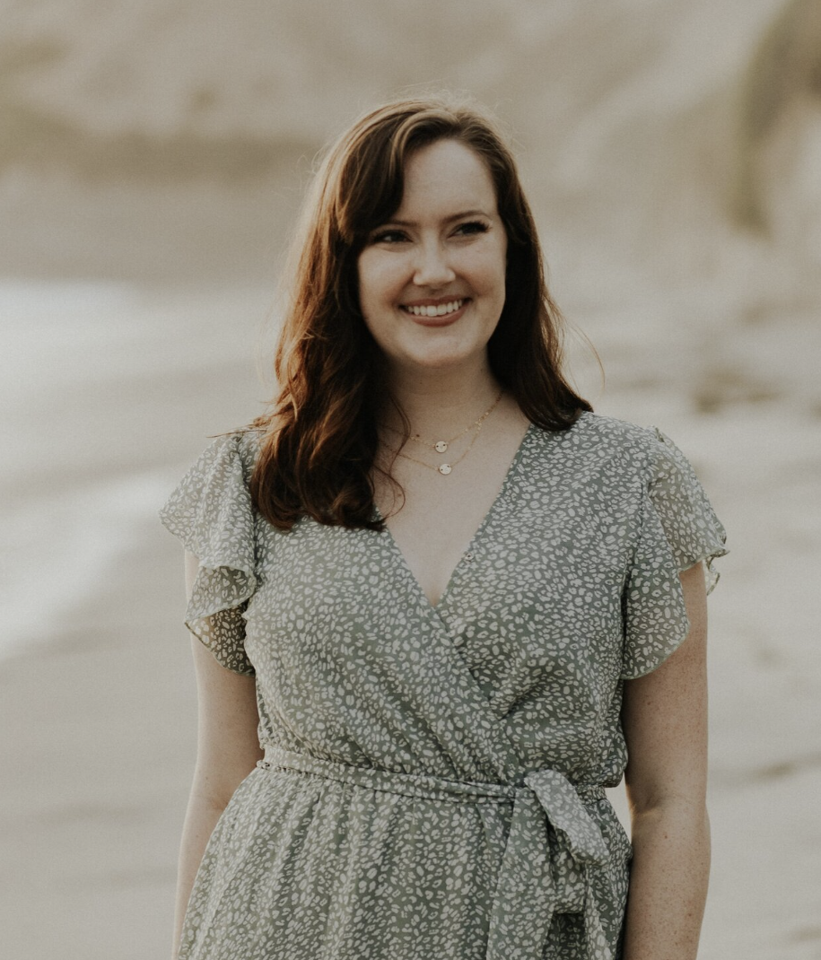 A woman with long brown hair wearing a gray patterned dress standing on a beach, smiling.