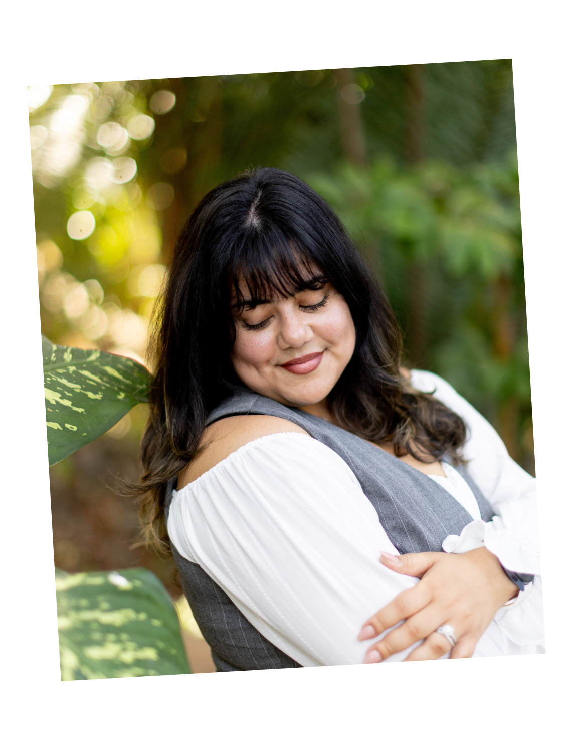 A woman with dark hair, closed eyes, and a gentle smile is hugging herself, standing outdoors with green foliage in the background.