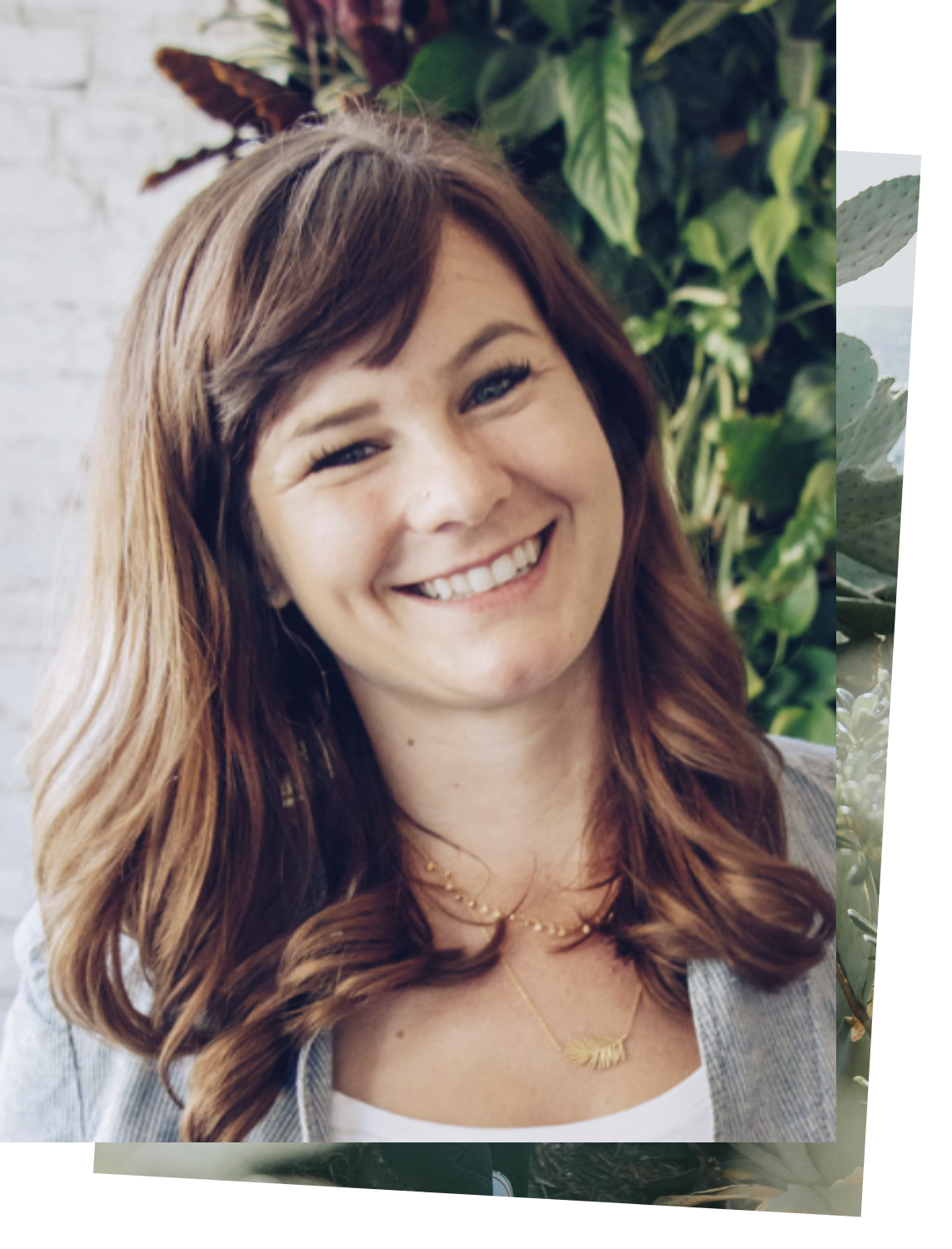 A woman with long wavy auburn hair smiling outdoors with green plants in the background.