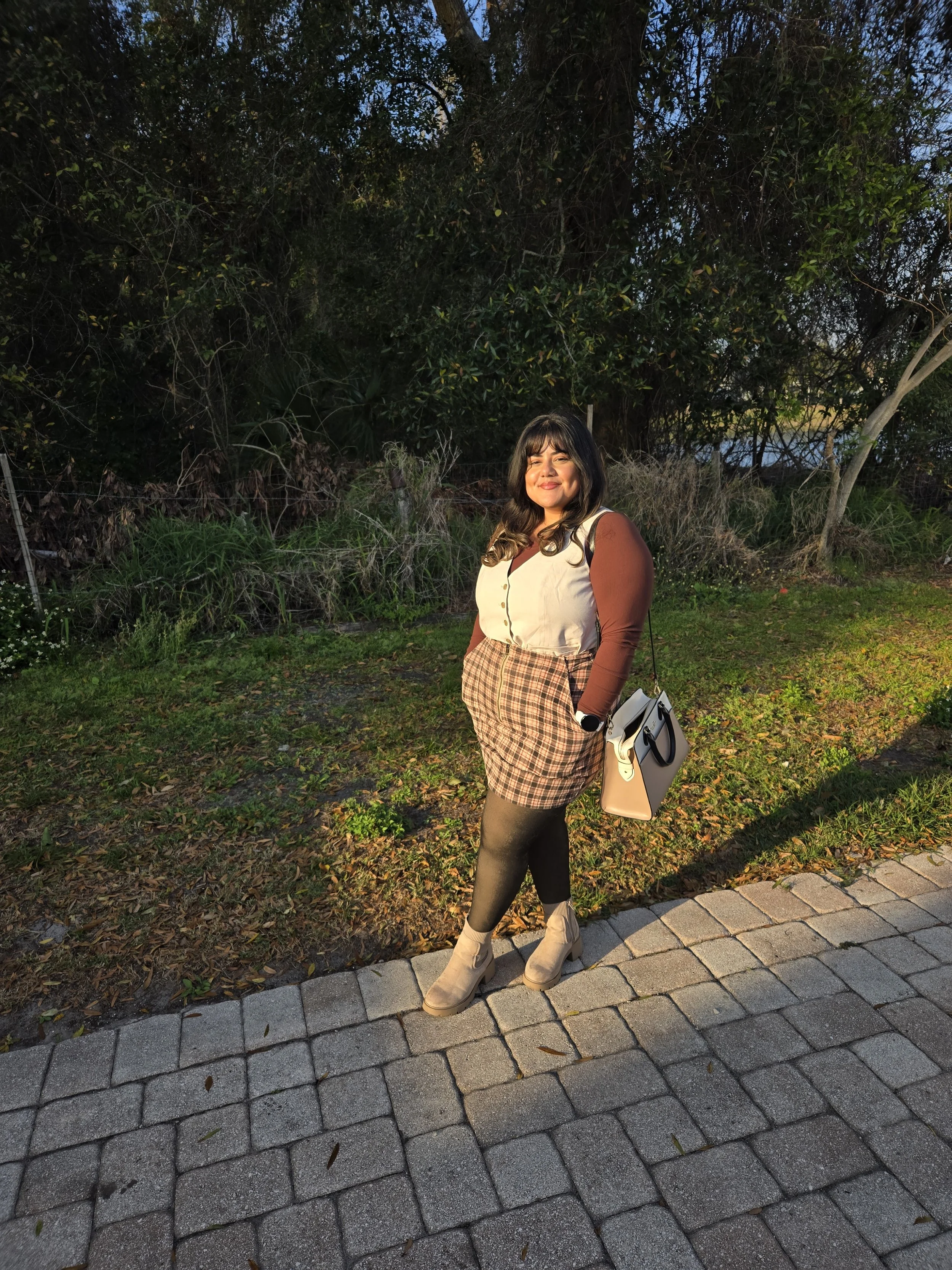 A woman standing on a park pathway at sunset, smiling at the camera. She is wearing a beige and brown outfit with checkered shorts, a beige vest, long brown sleeves, black tights, and beige boots. She has dark hair with bangs and carries a white handbag.