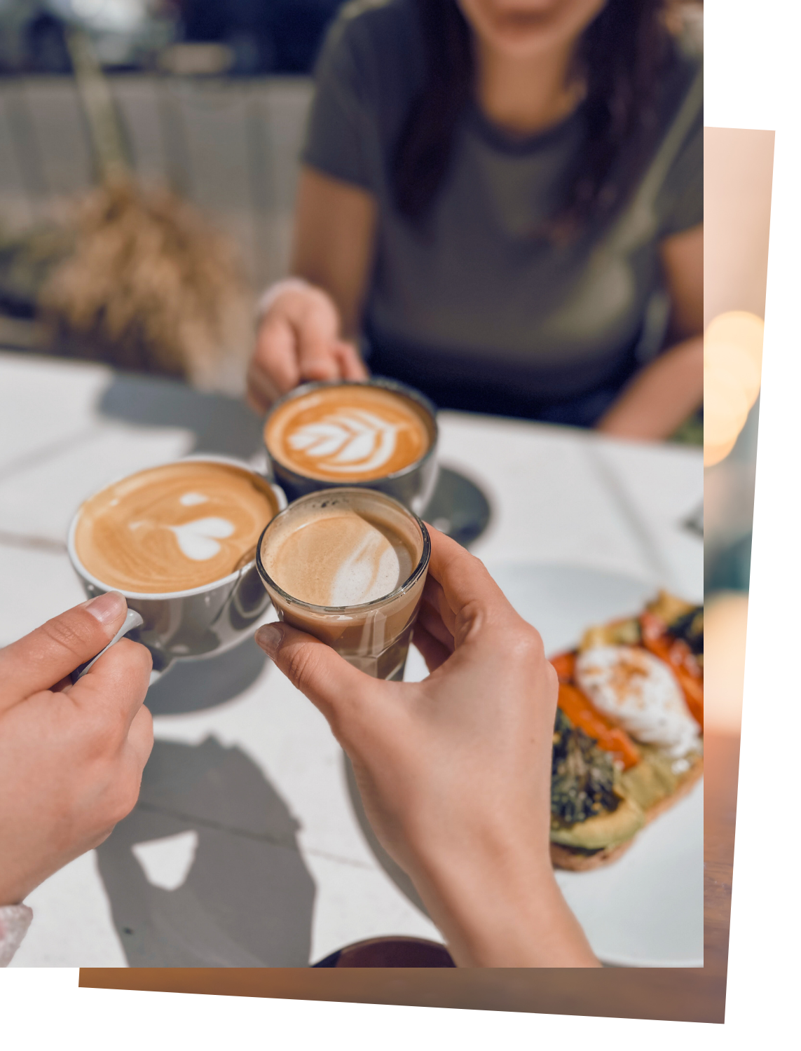 People raising cups of coffee in a toast at a table with food, including a sandwich topped with vegetables and a poached egg.