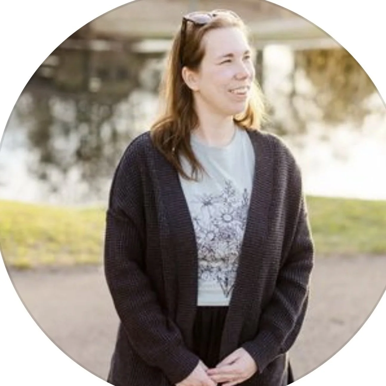 A woman with long brown hair wearing glasses on her head, a light gray T-shirt with a floral design, and a dark cardigan, standing outdoors near a body of water with trees in the background.