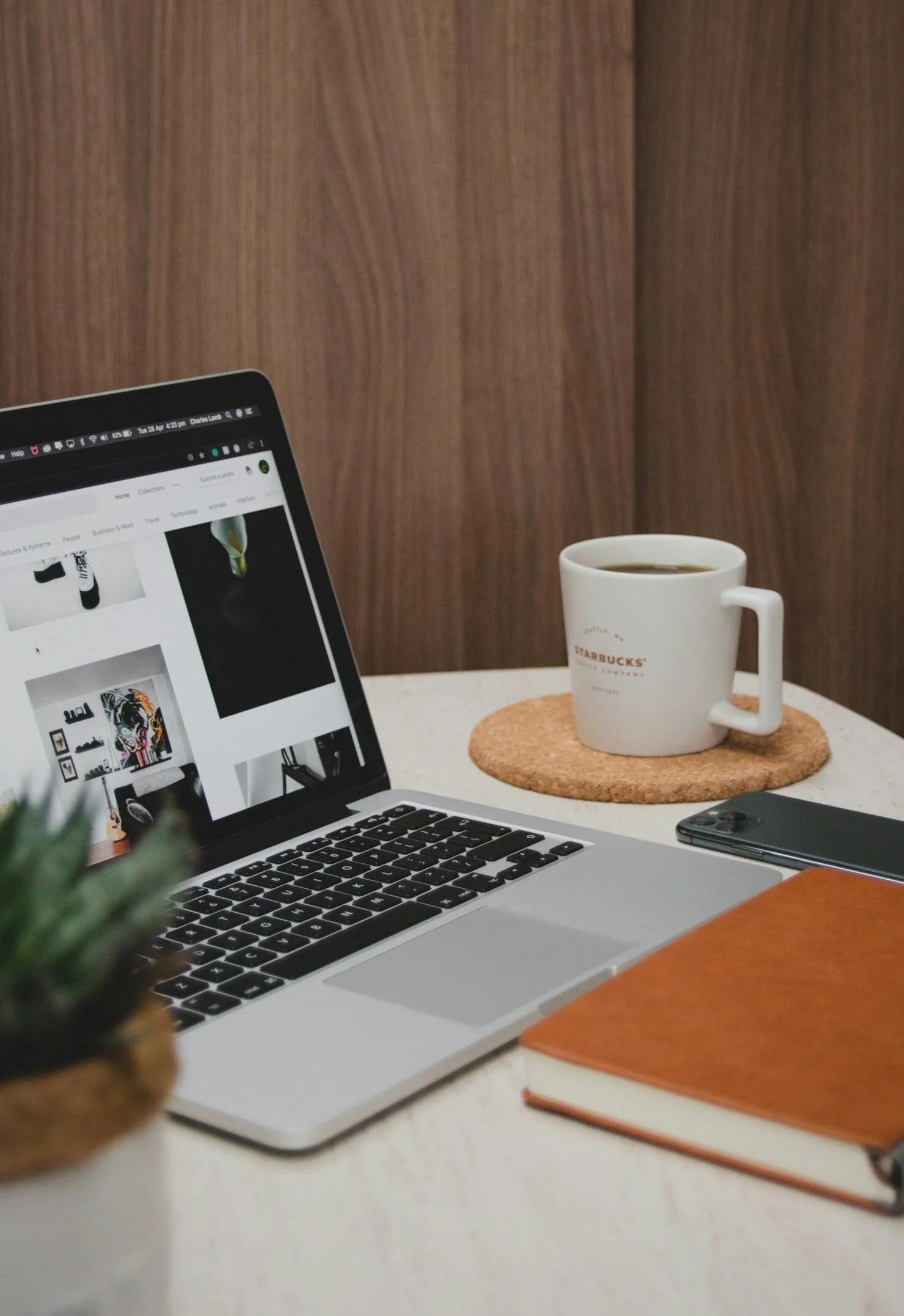 A workspace with a MacBook displaying a website, a cup of coffee on a cork coaster, a smartphone, a brown notebook, and a small potted plant on a white desk. A wooden wall is in the background.