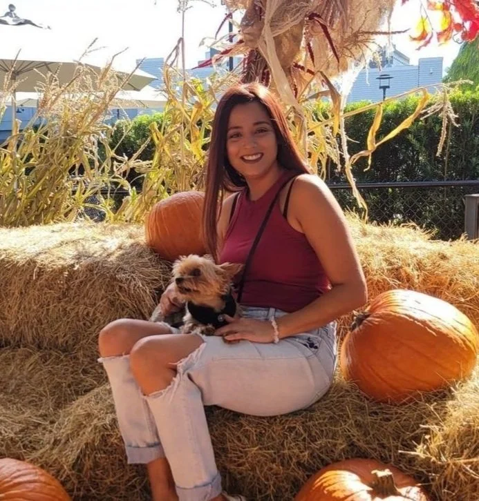 A young woman with long dark hair, wearing a maroon tank top and ripped light jeans, is sitting on hay bales with pumpkins around her. She is smiling and holding a small dog, with more pumpkins and dried corn stalks in the background.