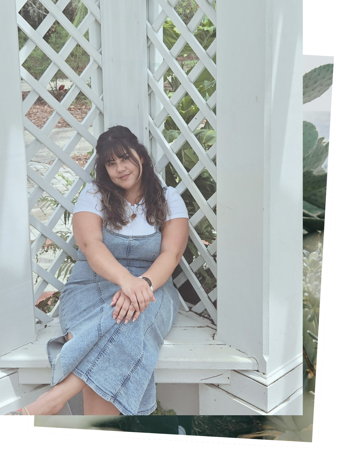 A woman sitting on a white wooden bench in front of a white lattice fence, surrounded by greenery and cacti.