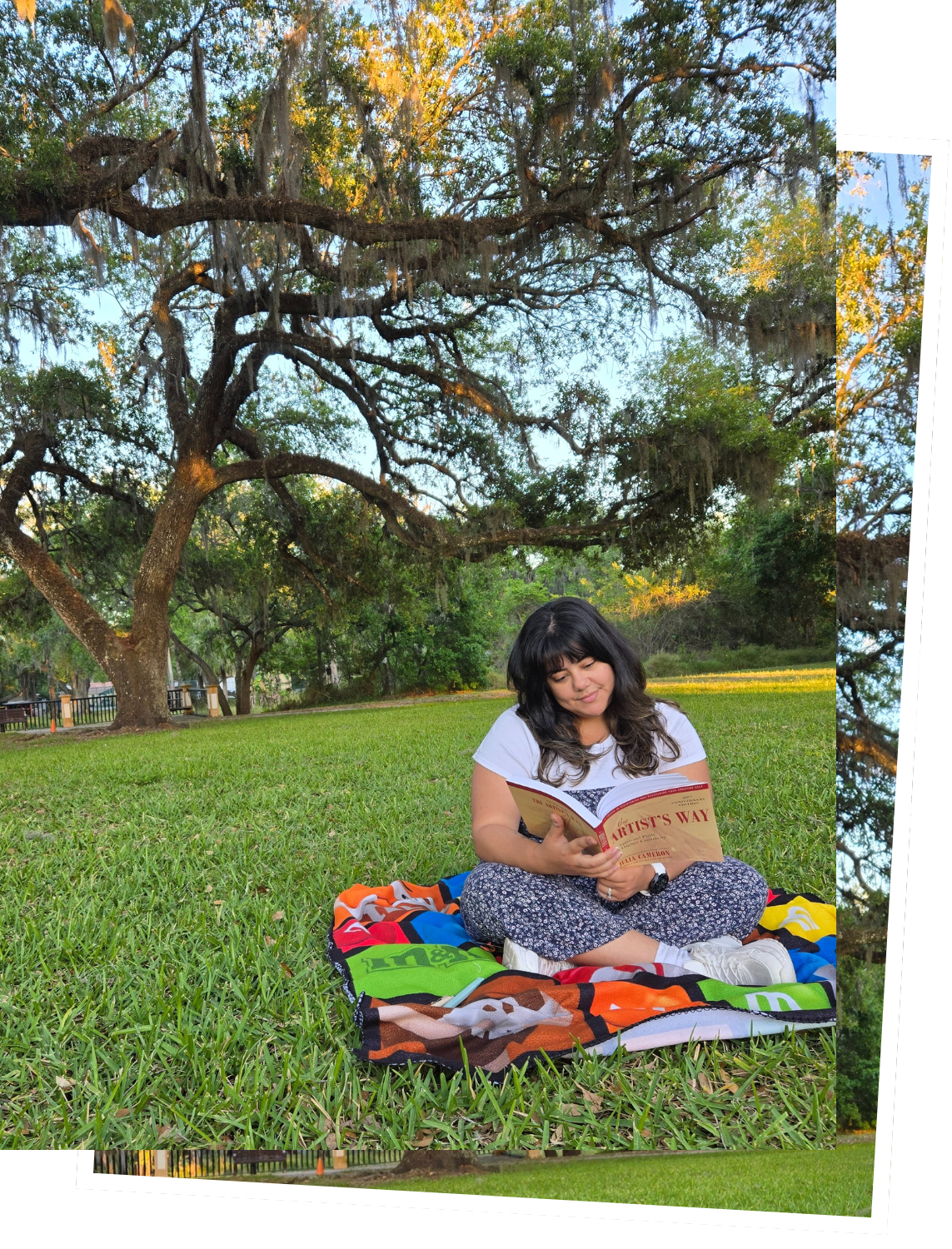 A young woman sitting cross-legged on a colorful blanket on the grass, reading a book titled 'The Artist's Way' in a park with large trees and green foliage in the background.