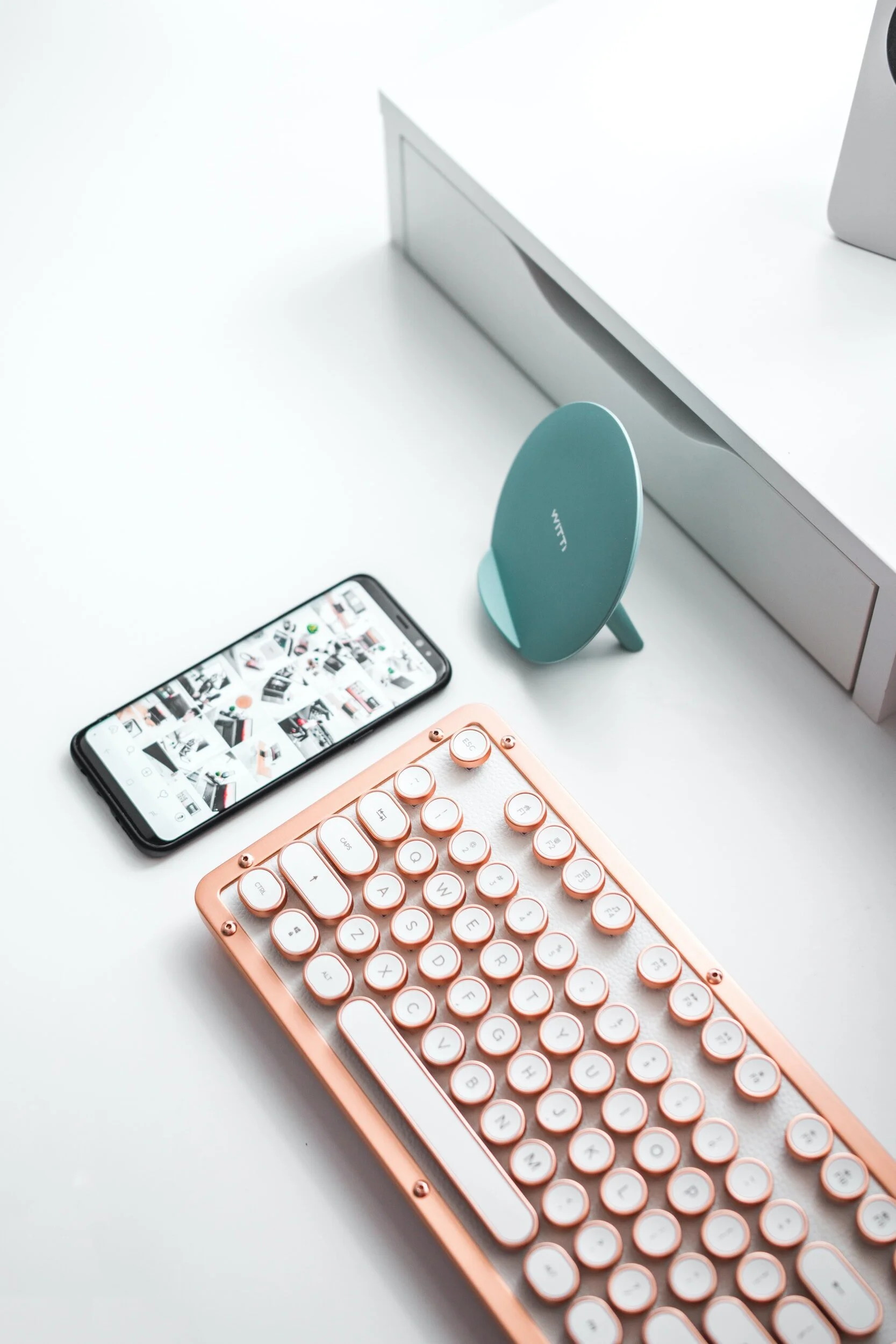 A pink wireless keyboard, a smartphone displaying a grid of images, a blue wireless charger, and a white box on a white desk.