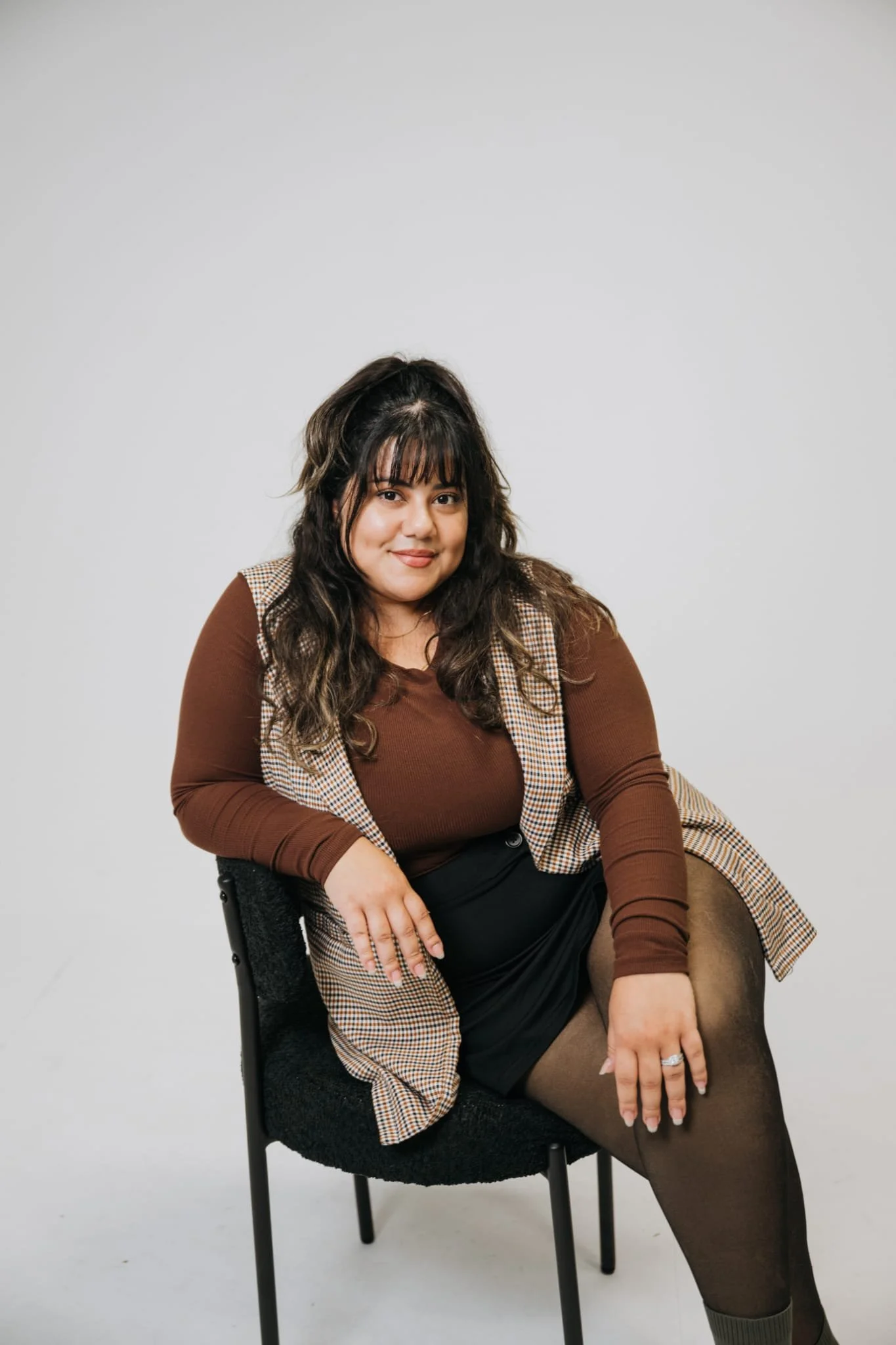 A woman with dark hair and bangs, wearing a brown long sleeve shirt, brown checked vest, black skirt, and patterned tights, sitting on a black chair against a plain white background, looking at the camera and smiling.