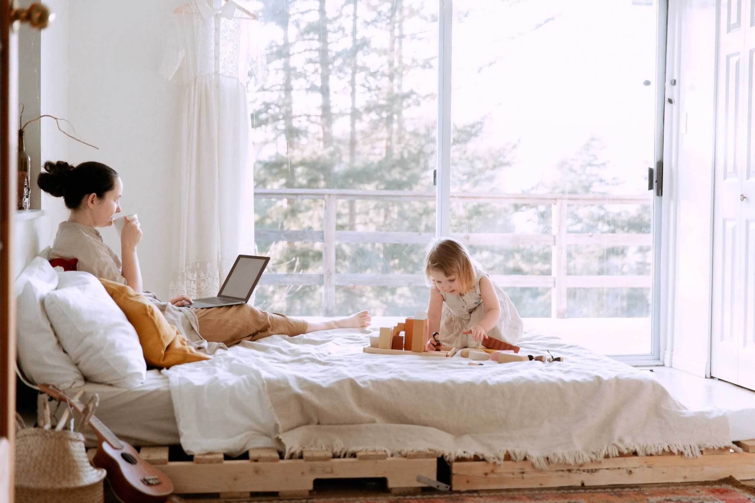 A woman sitting on a bed, drinking from a cup while using a laptop, as a young girl plays with wooden blocks on the bed in a bright room with large sliding glass doors and white curtains.