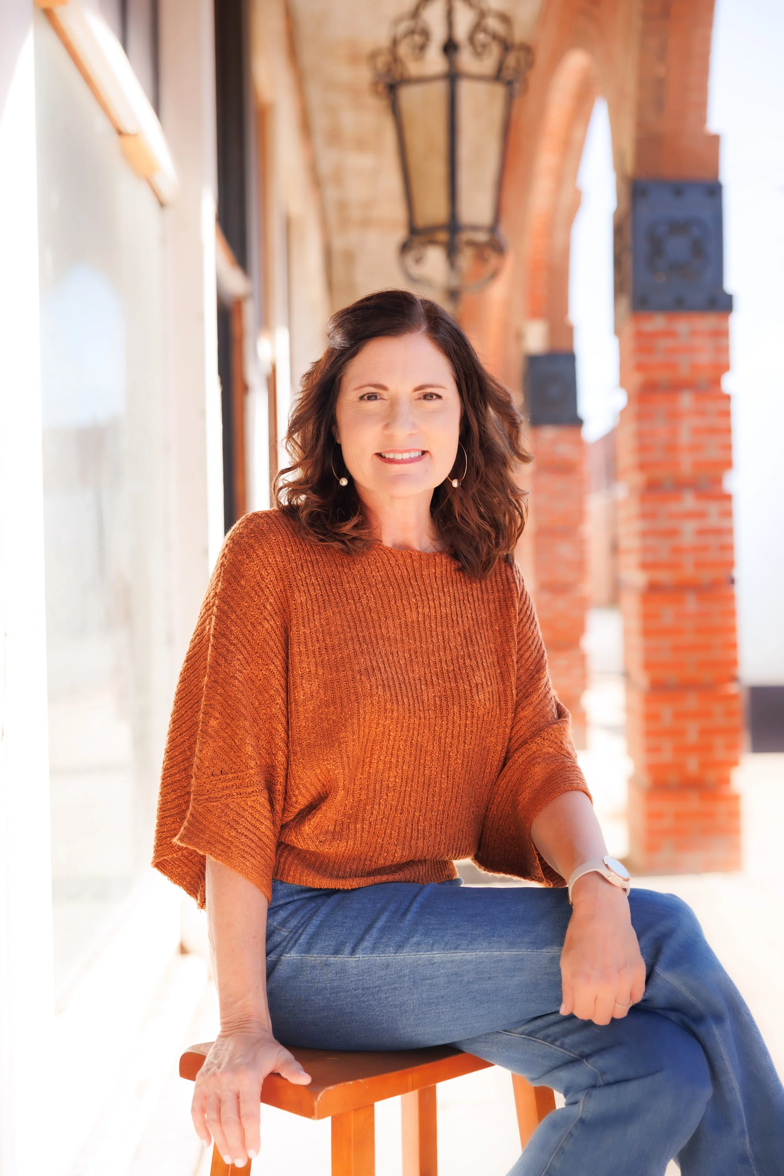 A woman with shoulder-length brown hair, wearing a black blouse and light gray pants, leaning against a tree outdoors with a blurred background of trees and grass, smiling at the camera.