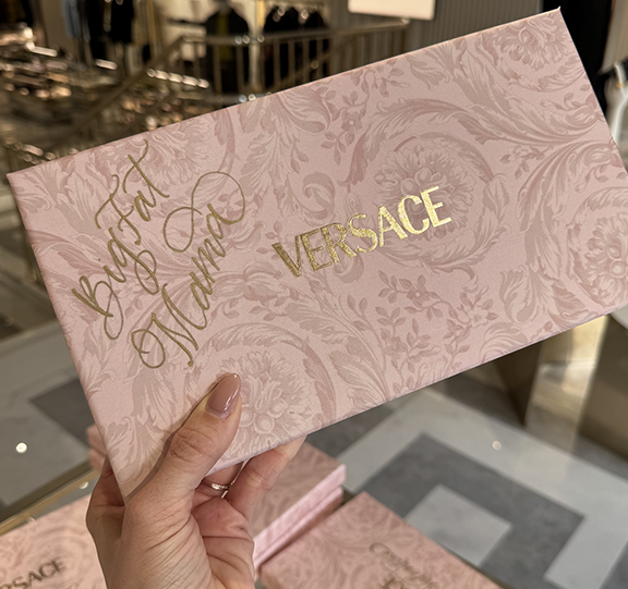A hand holding a pink patterned box with gold lettering that says "Beautiful and Wild" and the brand name "Versace" in gold.