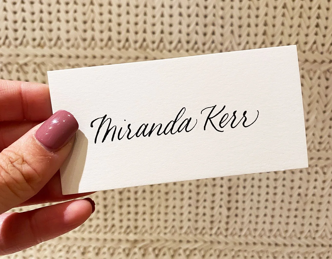 A person with painted nails holding a white card with the name 'Miranda Kerr' written in elegant black script. The background features a textured beige surface.