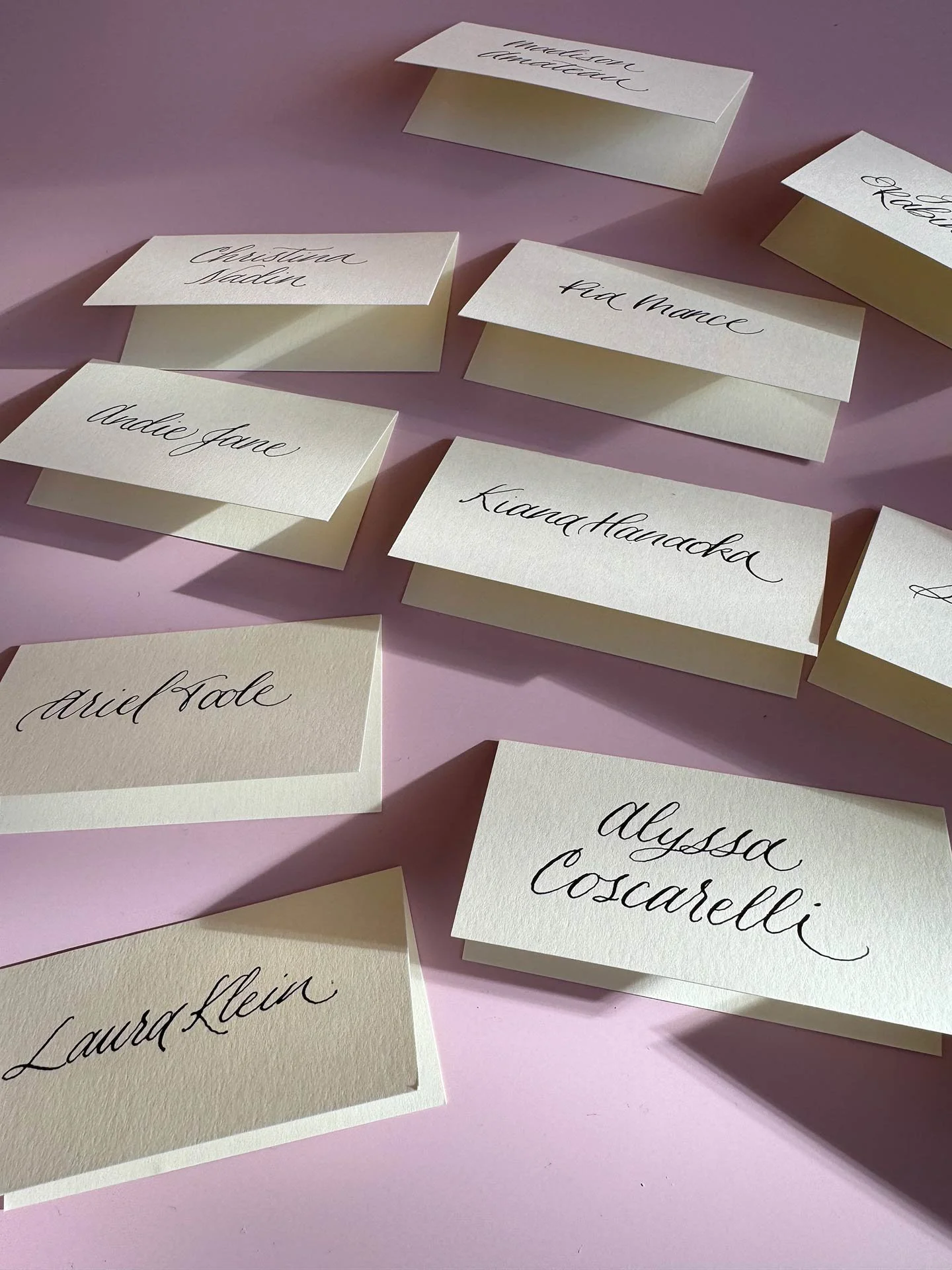 Collection of cream-colored place cards with handwritten names in black cursive, arranged on a pink surface.