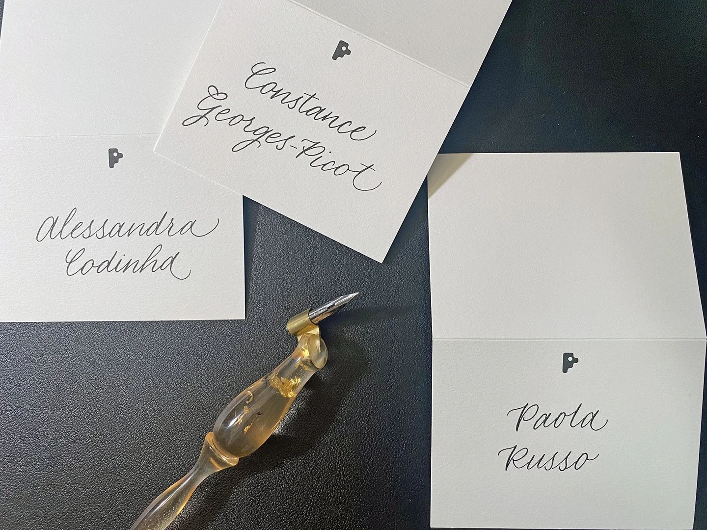 Three white cards with handwritten calligraphy names lie on a black surface, one card is partially on top of another, and there is a decorative gold pen with a glass body also on the black surface.
