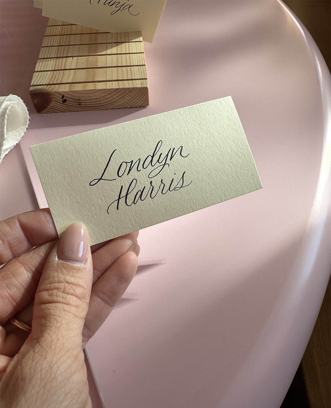 Hand holding a pale yellow card with the name "Londyn Harris" written in cursive, on a pink surface with a small wooden block and another card in the background.