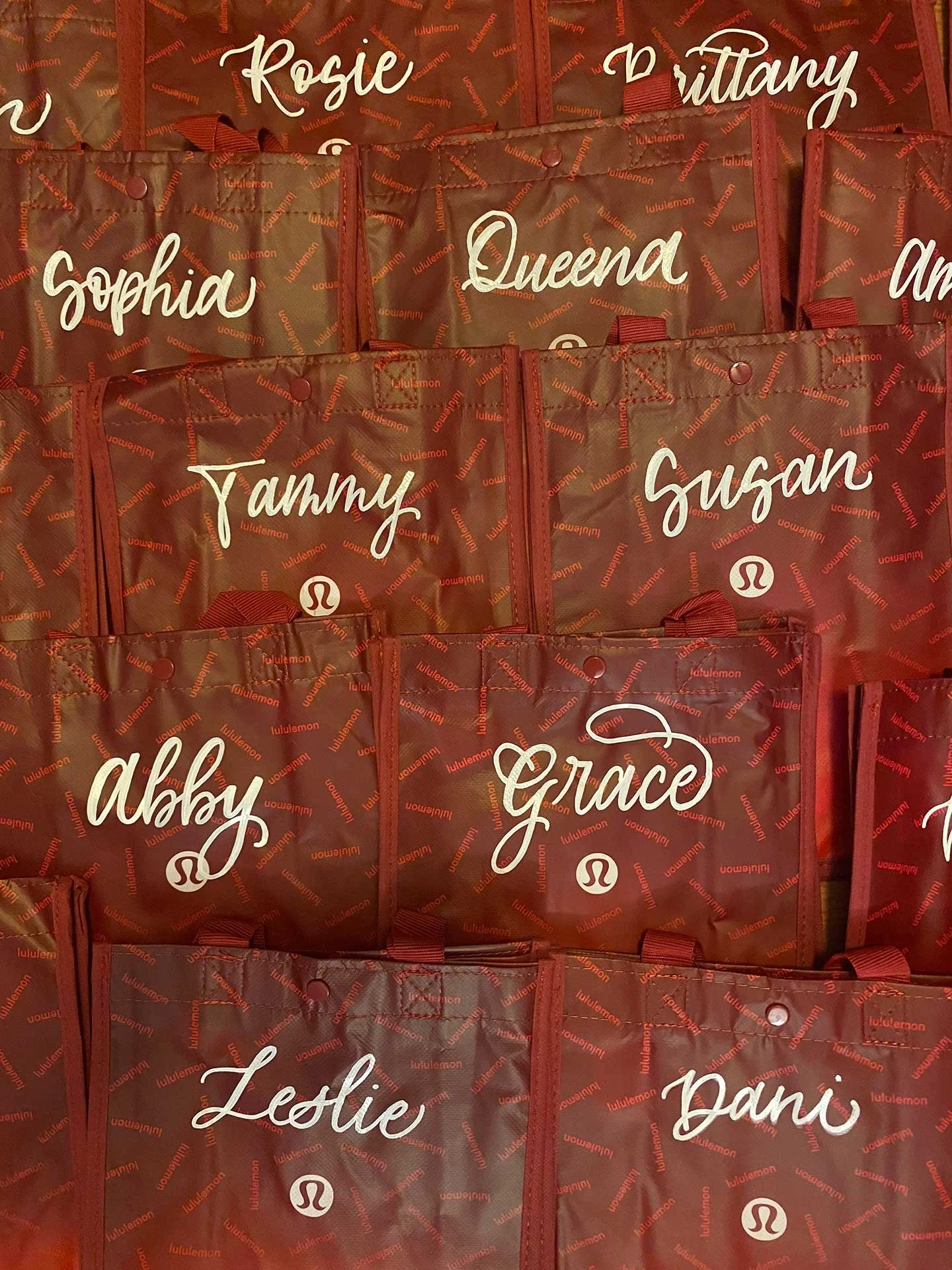 Red tote bags with white cursive names and the lululemon logo, including Rosie, Brittany, Sophia, Queena, Amy, Susan, Abby, Grace, Leslie, and Dani.