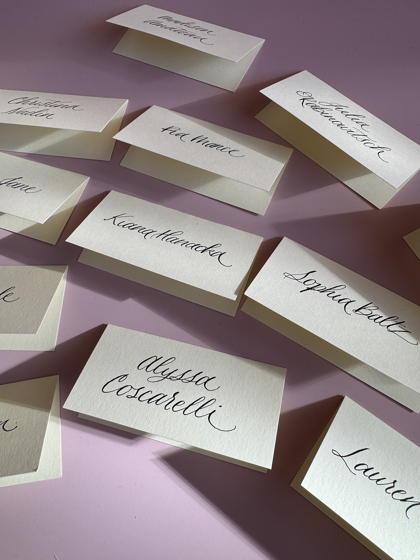 Several cream-colored name cards with handwritten calligraphy, featuring names like 'Alyssa Casarelli,' 'Kait Handwerker,' 'Lydia ballz,' 'Lauren,' 'Kristen Nadel,' and 'Kate Nance,' spread out on a pink surface.