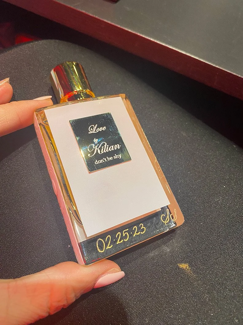 A perfume bottle with a gold cap and a label that says 'Love by Kilian don't be shy.' The bottle is on a dark surface, with the time 02:25:23 displayed at the bottom.