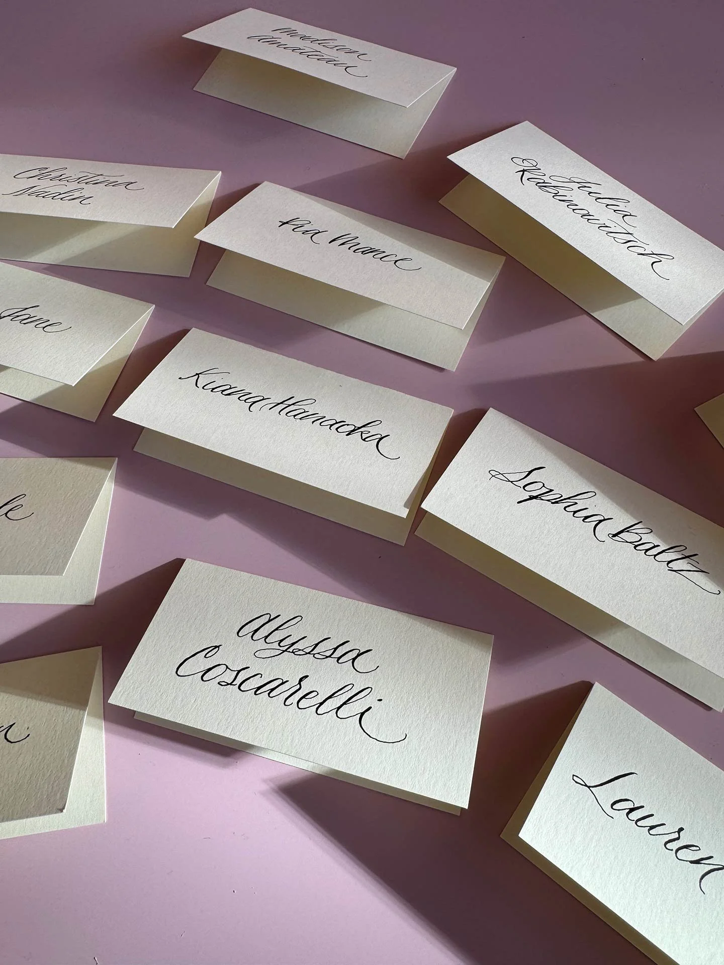 Collection of white name cards with cursive handwritten names, scattered on a pink surface.