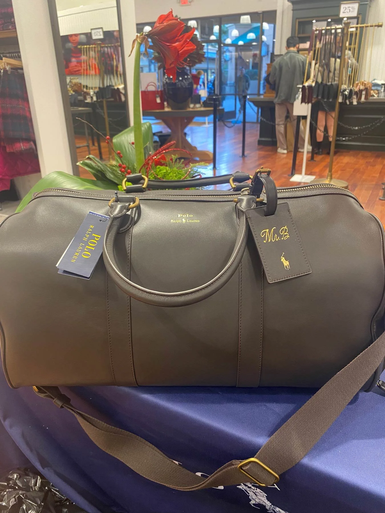 A gray Polo Ralph Lauren duffle bag with black handles and a brown adjustable shoulder strap is placed on a table inside a retail store. There is a red amaryllis flower with green leaves next to the bag. The background shows store displays, shelves w