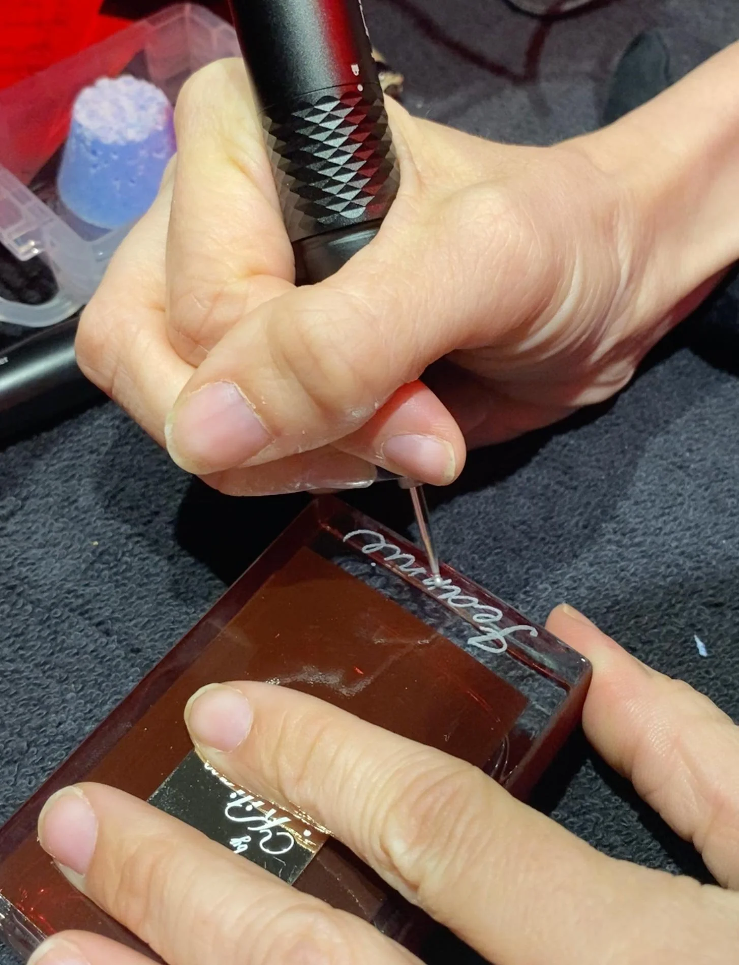 A person engraving cursive text onto a glass or acrylic object with a small engraving tool, while holding it steady with other fingers.