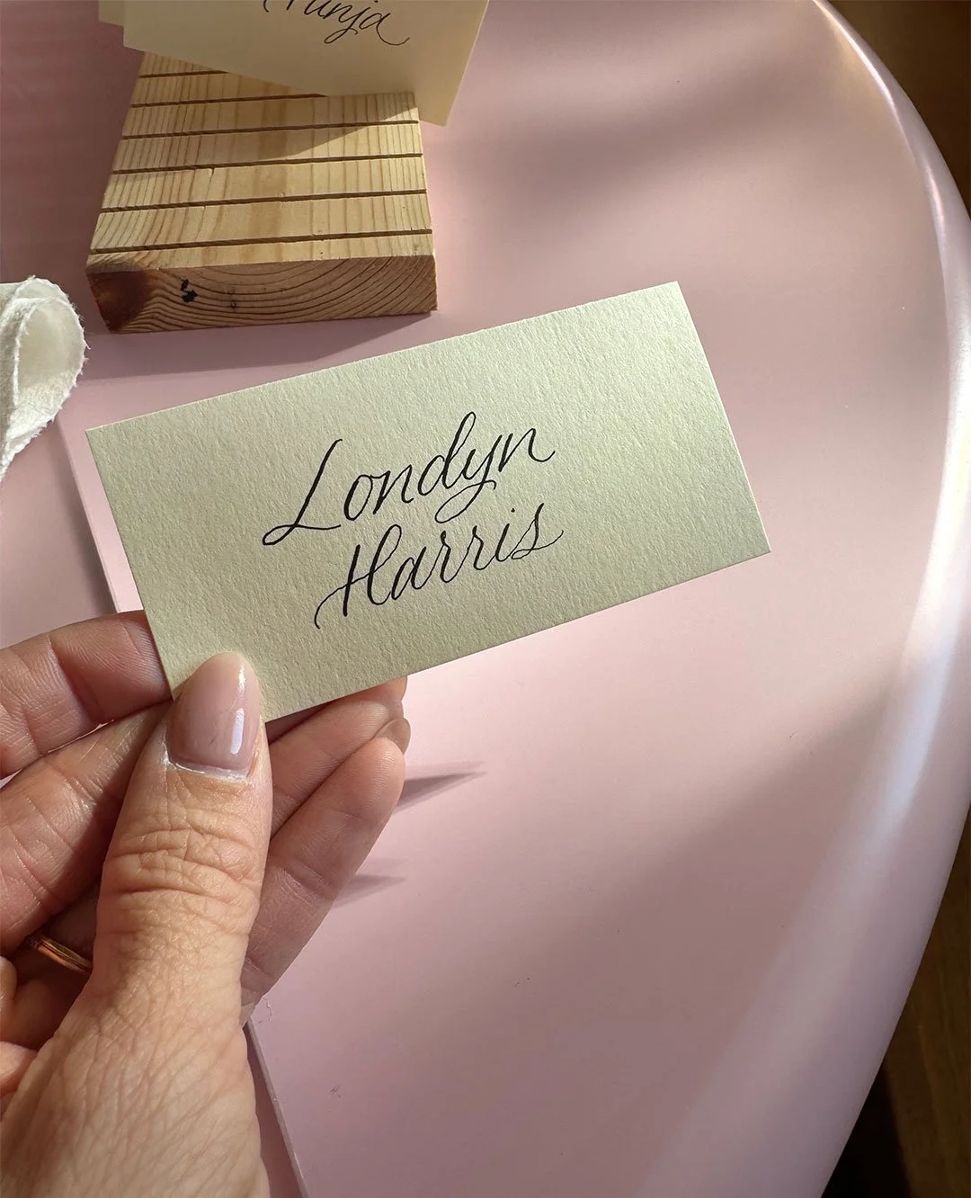 Hand holding a small note card with the name "Londyn Harris" written in elegant cursive, with a pink surface and a wooden block with similar notes in the background.
