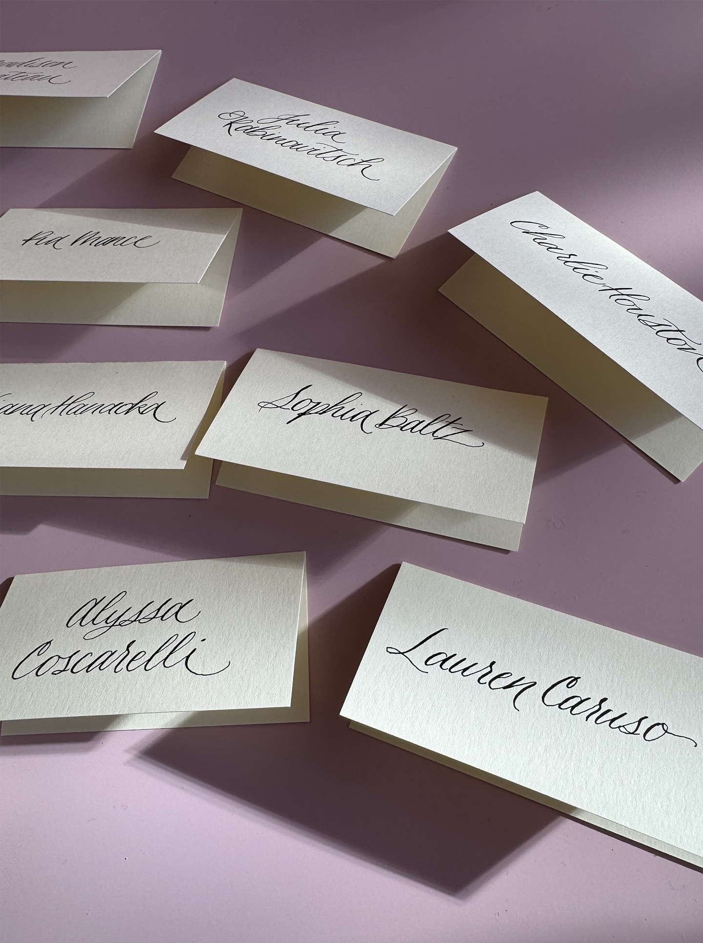 Several white place cards with handwritten names in black cursive font, scattered on a pink surface. Names visible include Lauren Consono, Alysaa Coscarelli, Sofia Baitz, Charlotte Houw, Flo Mace, and Olivia Handled.