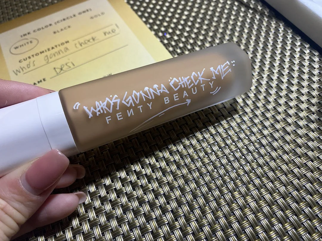A beige bottle of Fenty Beauty foundation with white handwritten text that says "Who's gonna check me". The bottle is held in a hand with a pink polished nail. In the background, there is a yellow note and a woven table surface.