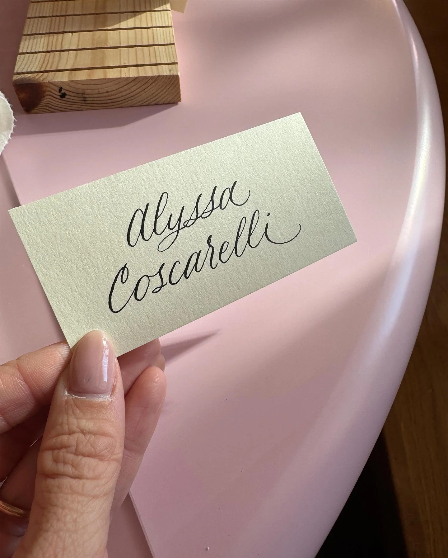 A hand holding a cream-colored card with the words 'alyssa Coscarelli' written in script, on a pink table with a wooden block nearby.
