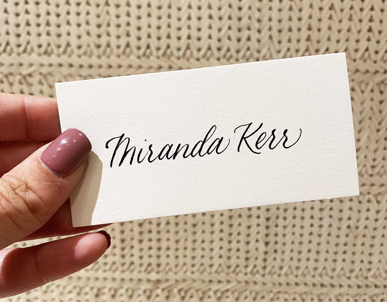 Close-up of a person's hand holding a white name tag with the name 'Miranda Kerr' written in elegant black script font, against a textured beige background.