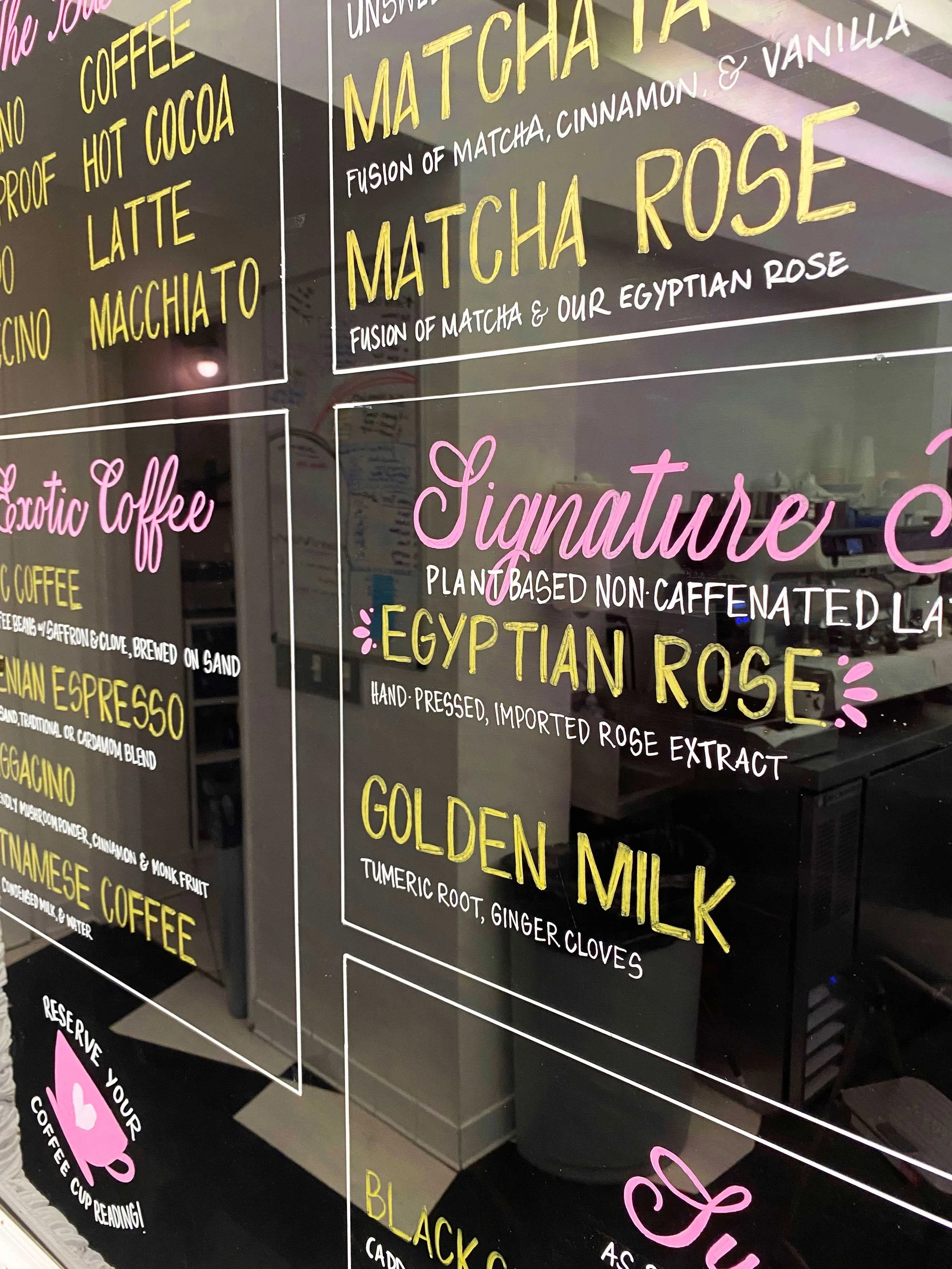 Close-up photograph of a colorful coffee shop menu on a black backlit board, featuring various coffee flavors and signature blends written in yellow, pink, and white chalk-like text.