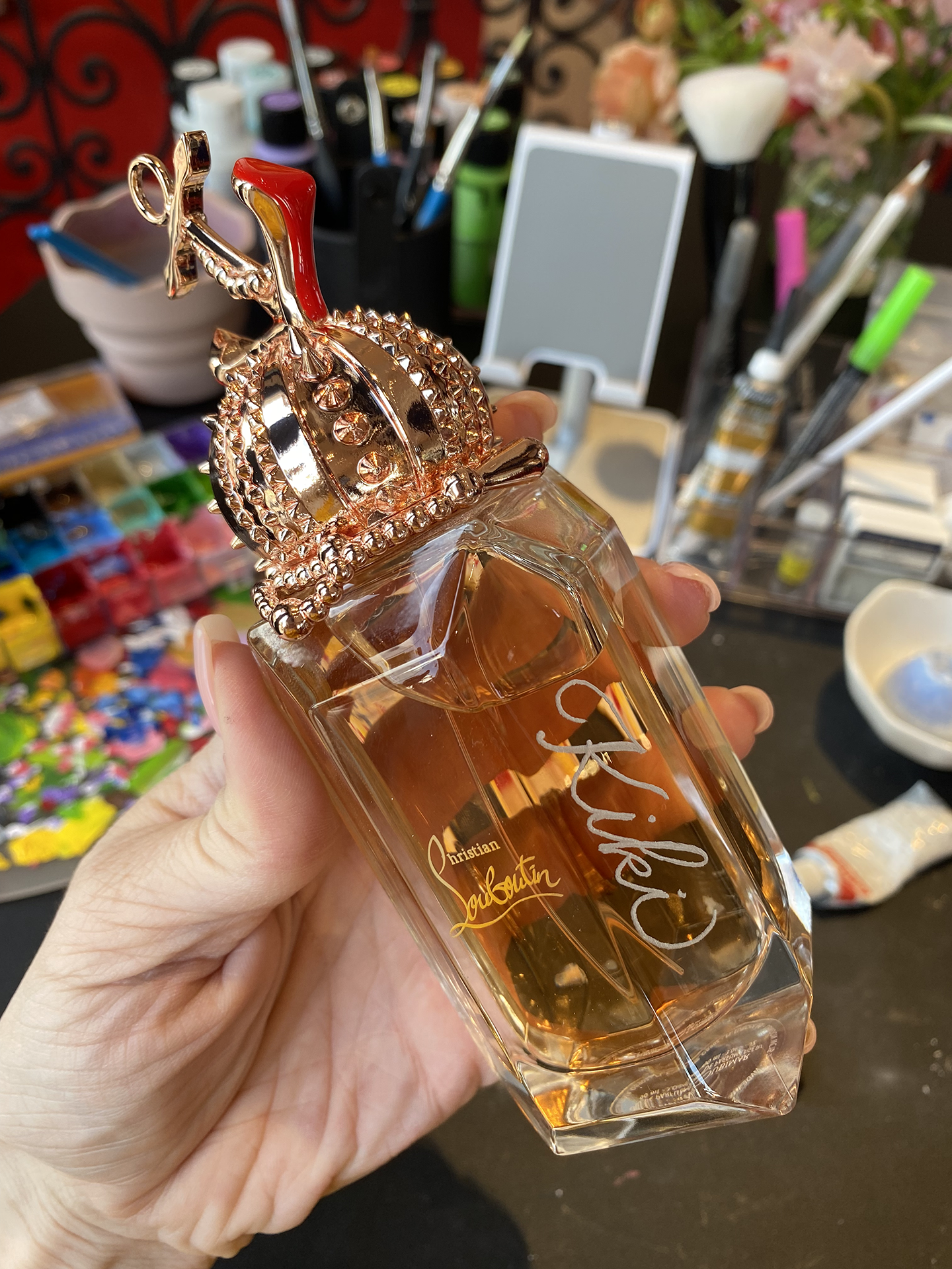 A Christian Dior J'adore perfume bottle with a gold crown top, held in a person's hand. Background includes makeup brushes, a mirror, and various beauty products.