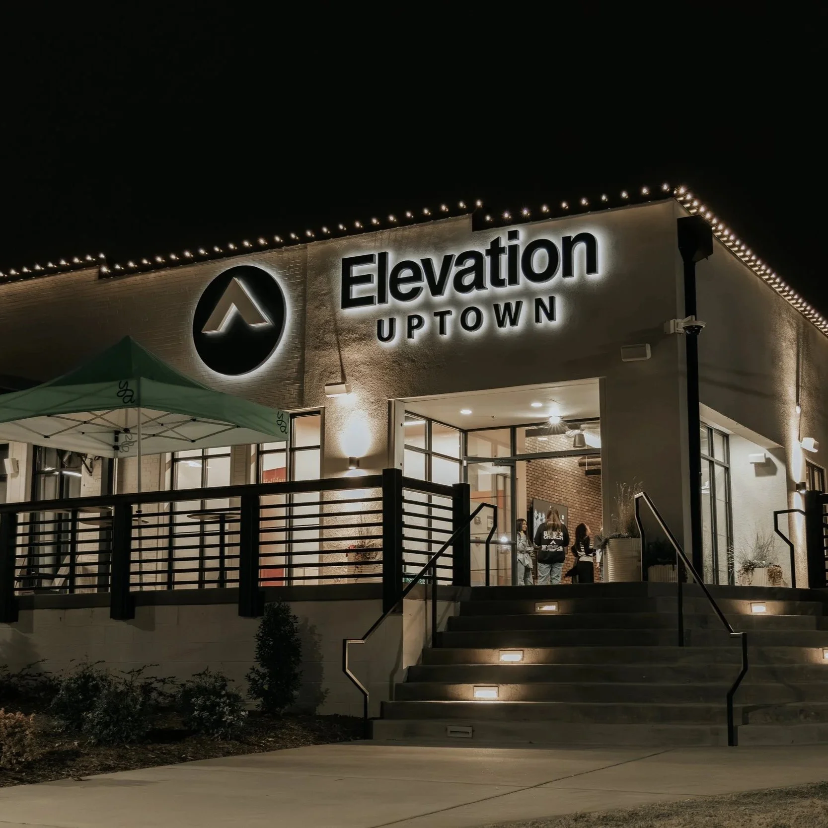 Night view of Elevation Uptown building illuminated with signage and string lights, with stairs and a small outdoor patio area.