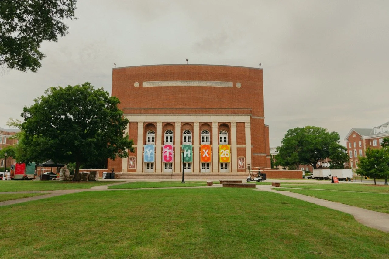 A large brick building with tall windows and decorative columns, with colorful banners and signs promoting an event called 'CHX 26' on a grassy area with walkways and trees in the foreground.