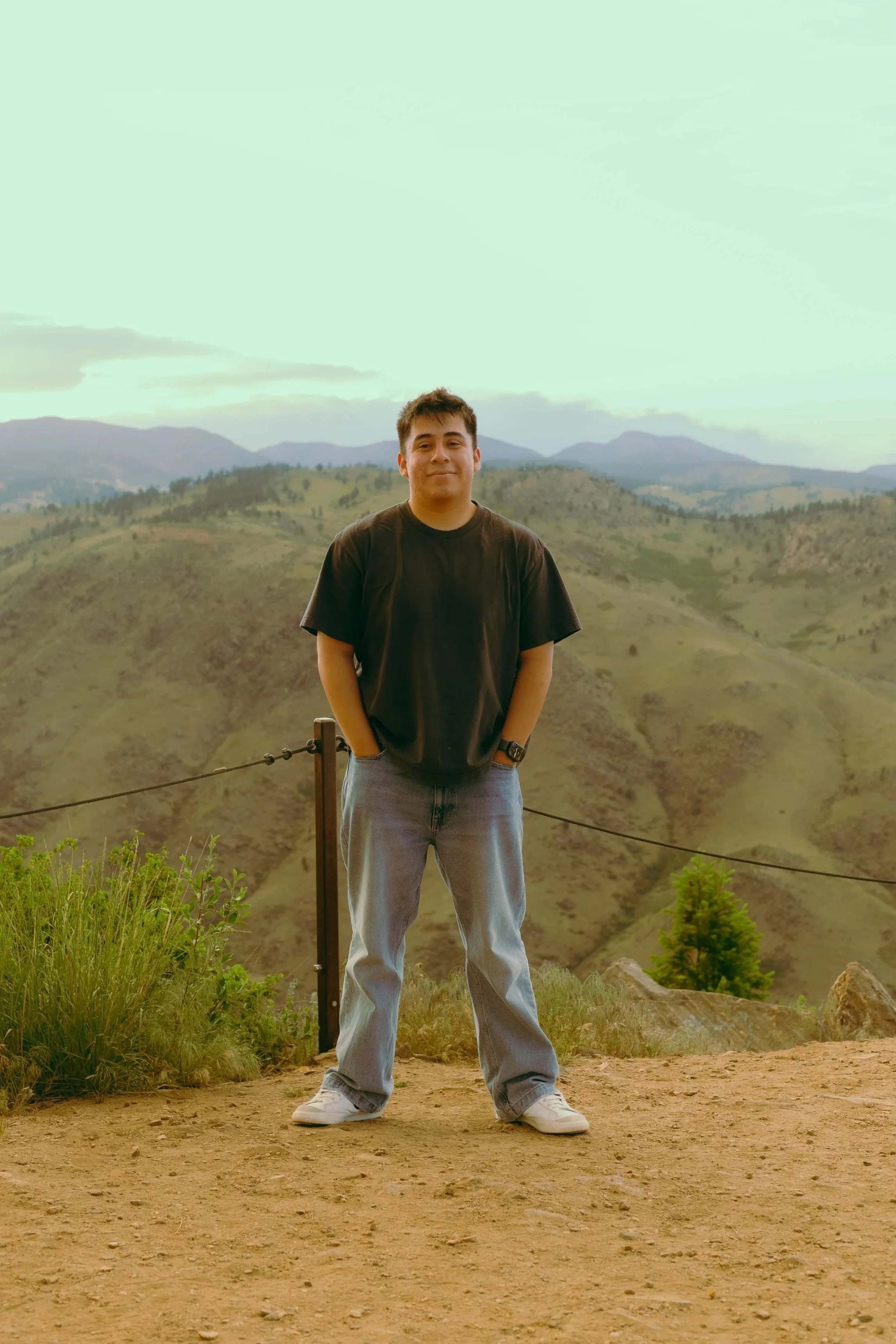 A young man standing outdoors with mountains in the background, wearing a black t-shirt, light blue jeans, and white sneakers.