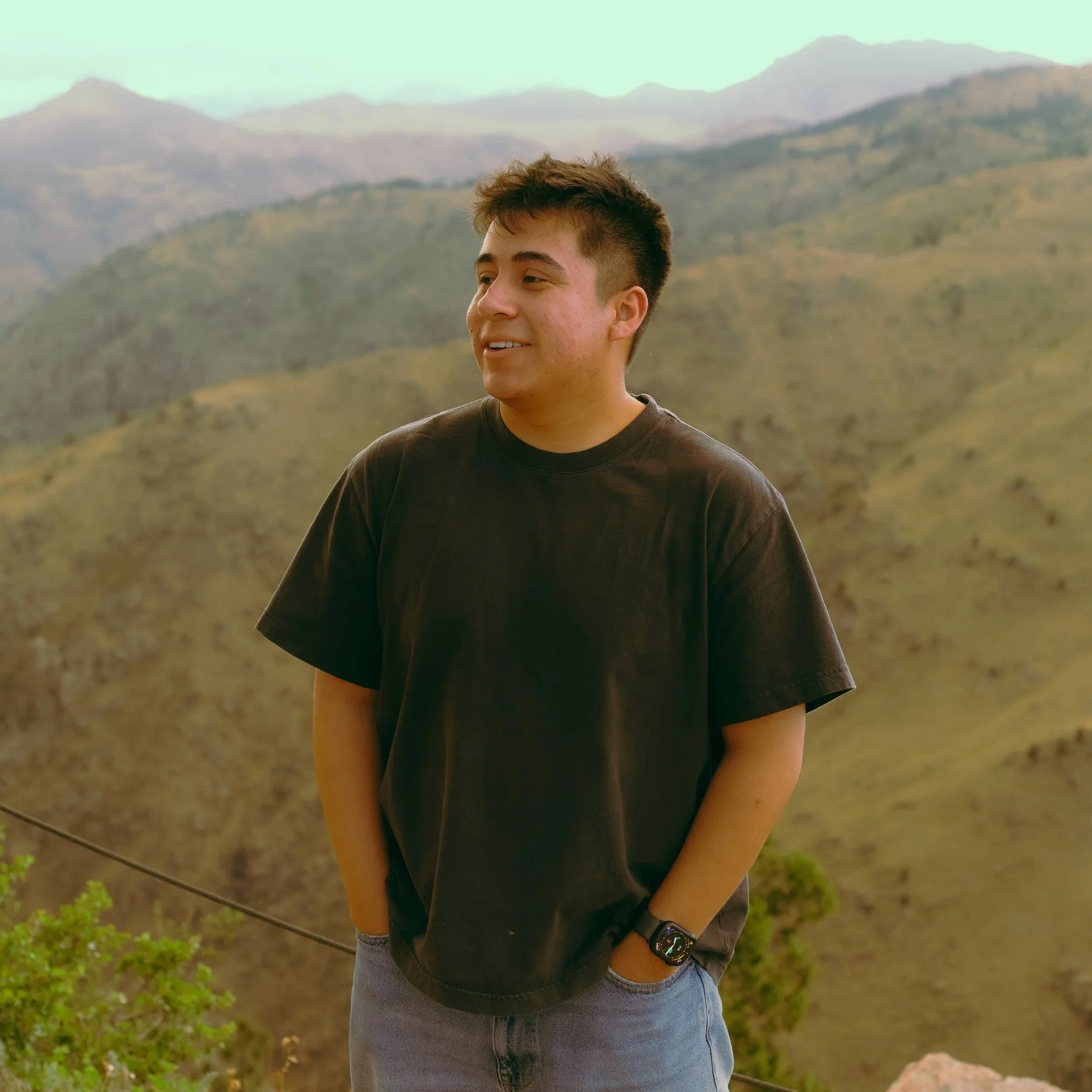 A young man with short dark hair, wearing a dark t-shirt and a watch on his left wrist, standing outdoors with a mountainous landscape in the background.