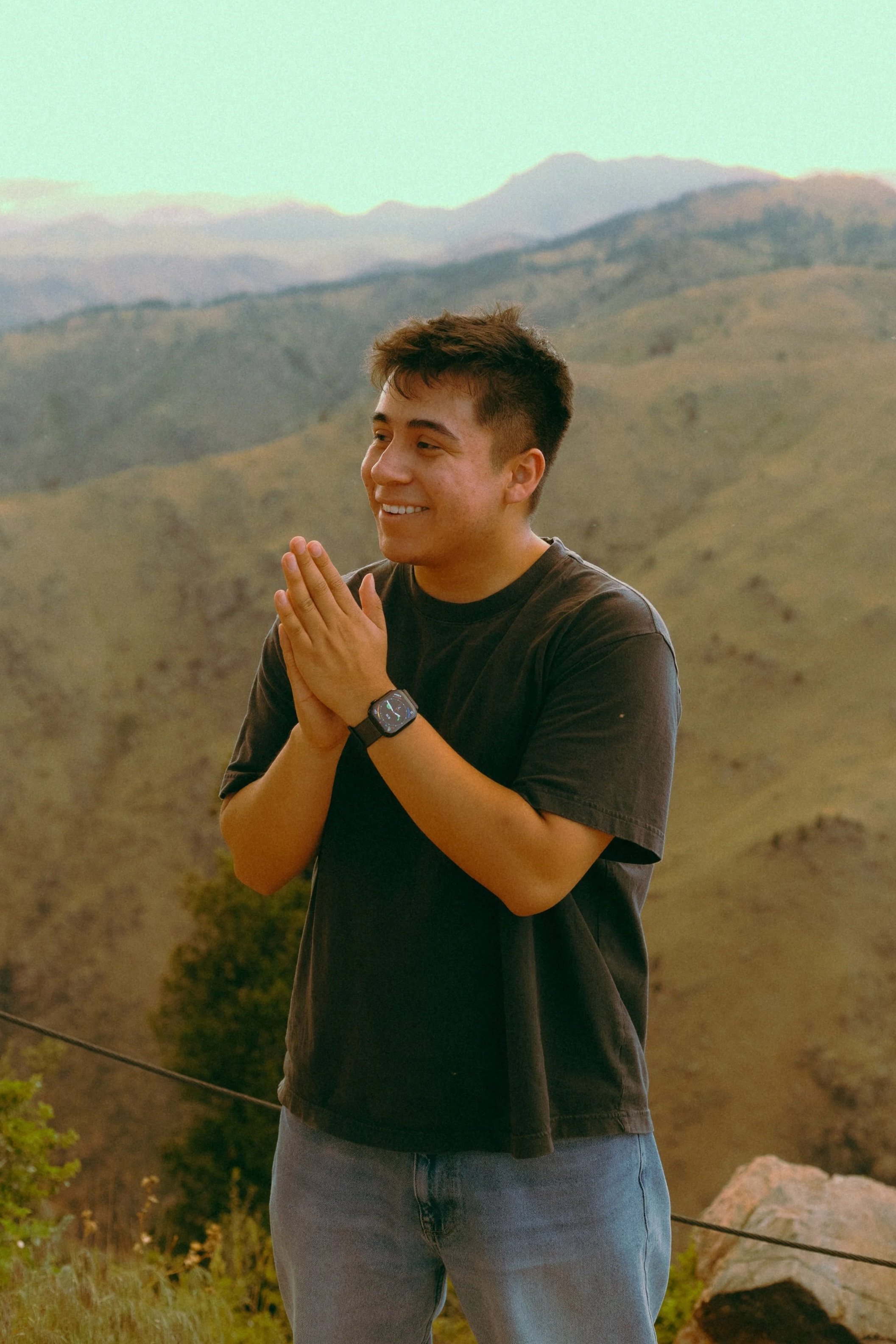 Young man smiling outdoors at sunset, hands together in a prayer or greeting gesture, in a mountainous landscape.