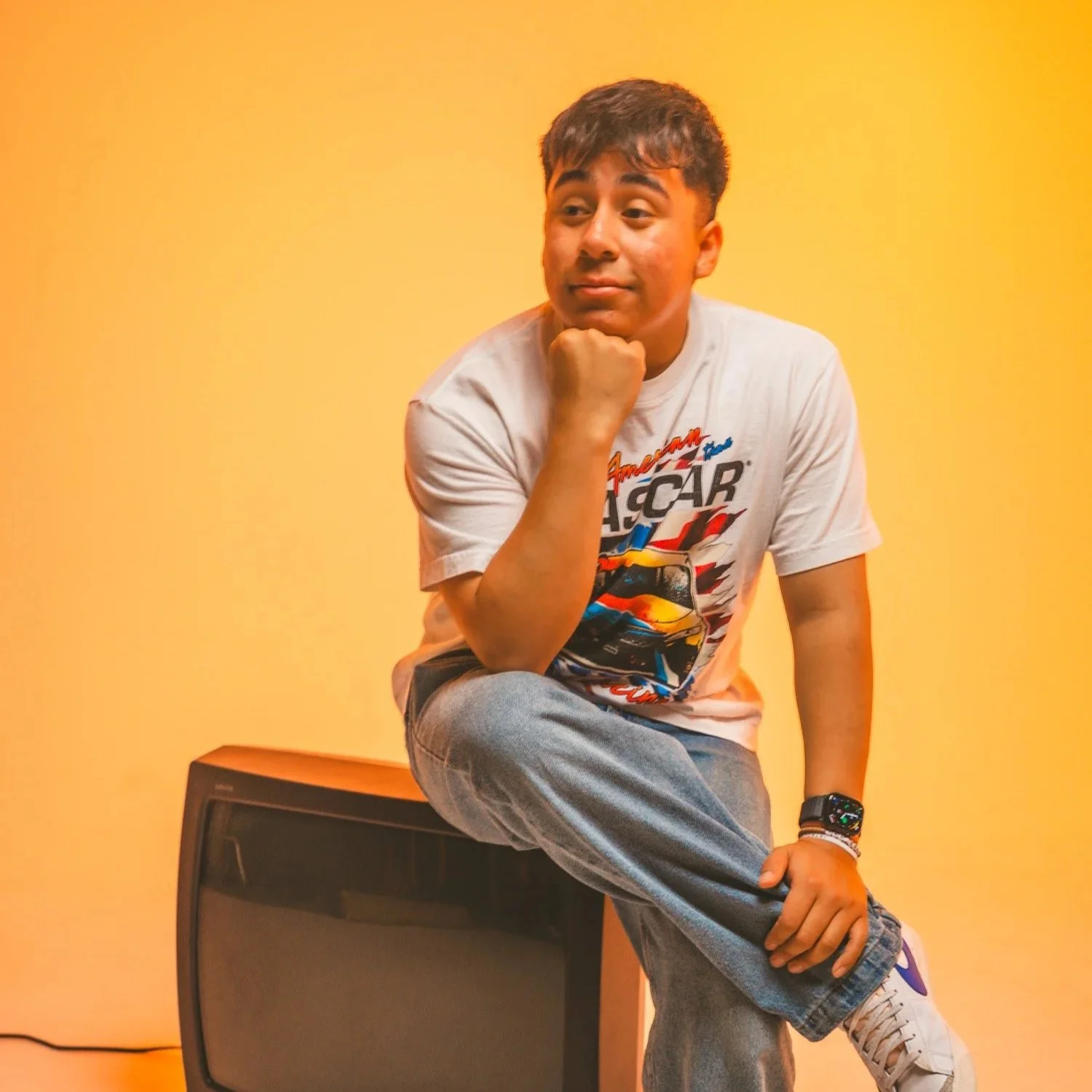 A young man sitting on a vintage television set against a yellow background, wearing a white NASCAR T-shirt, gray jeans, and a smartwatch, with one arm resting on the knee and his chin resting on his hand.