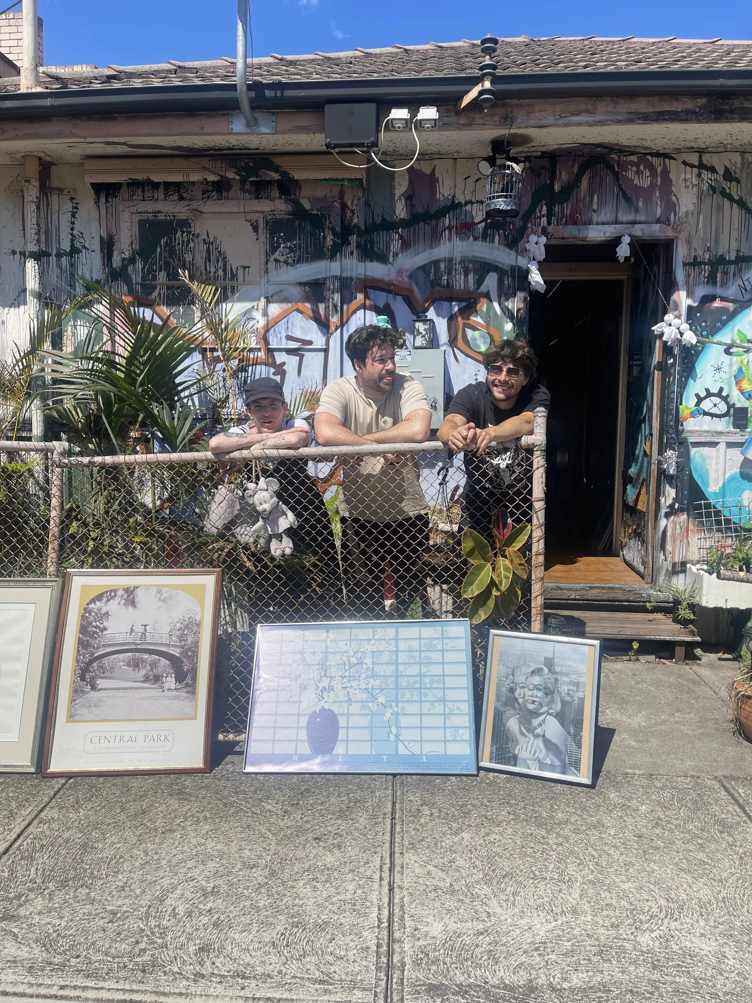Three people standing behind a chain-link fence in front of a graffiti-covered building, with artwork and photos displayed on the ground in front, and a clear blue sky overhead.